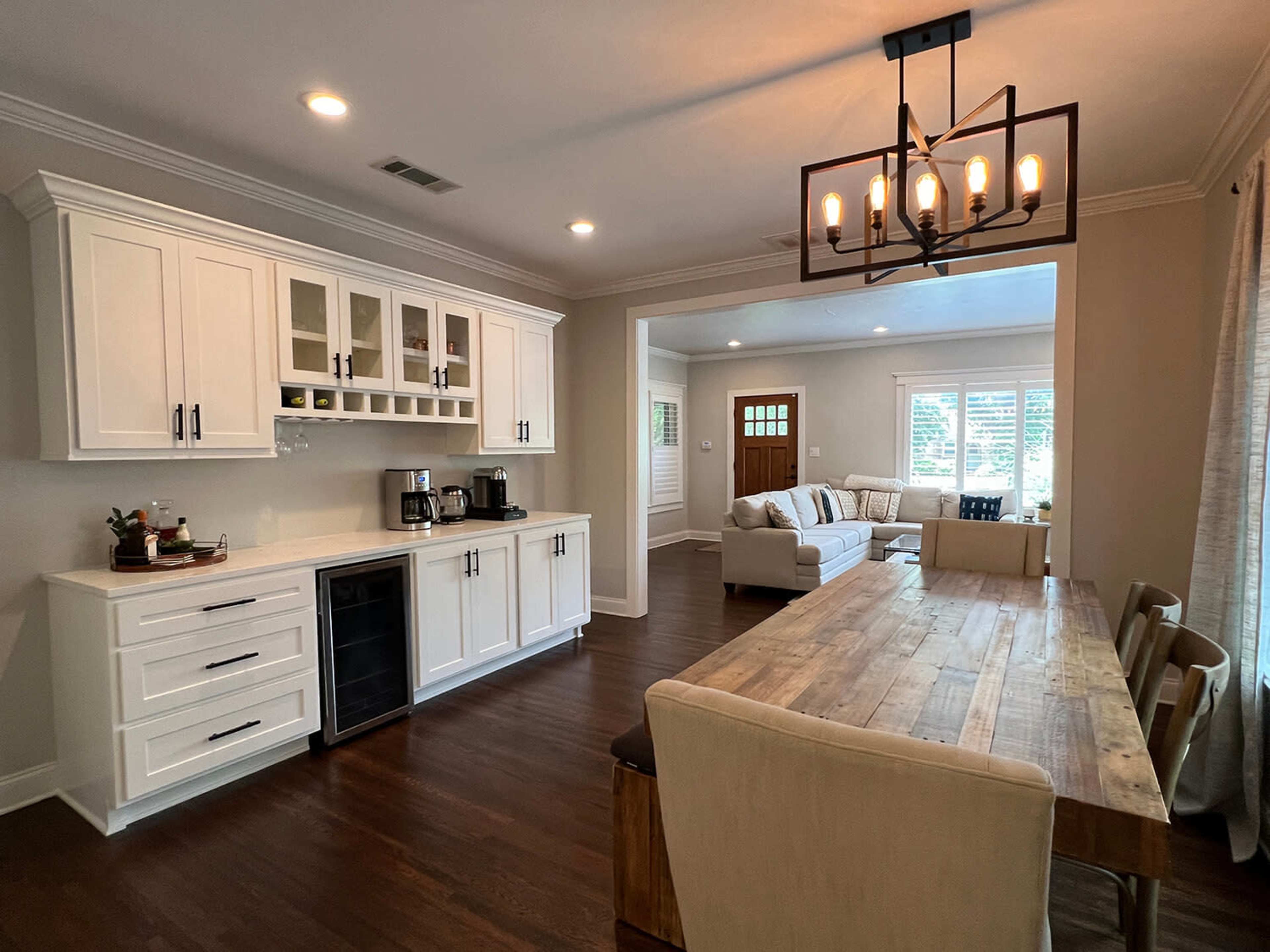 The image depicts a modern kitchen and dining area featuring white cabinetry, a wooden dining table, and a living room visible in the background.