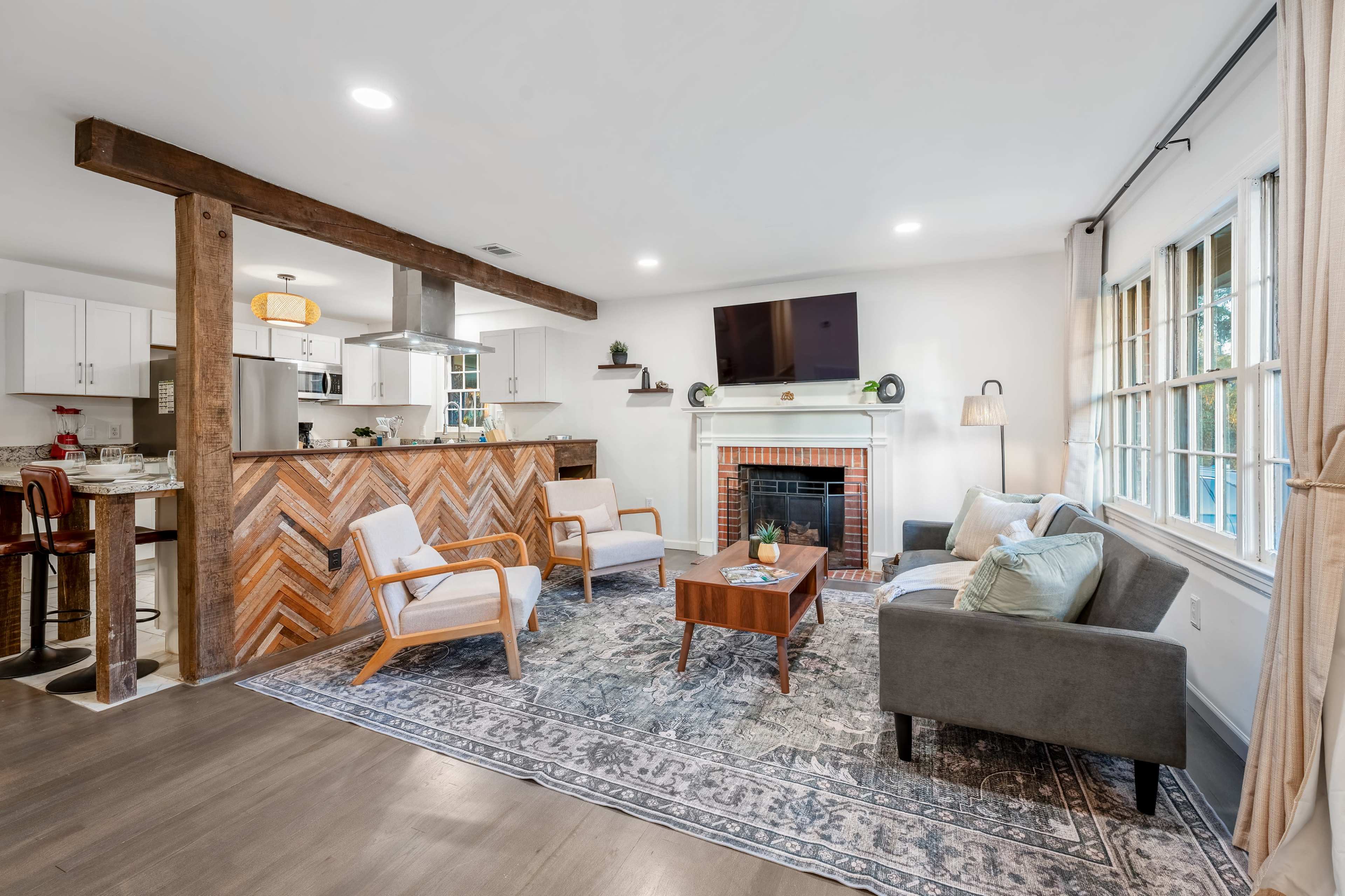 The image shows a modern living room with a fireplace, hardwood floors, and a combination of white and wooden furnishings, featuring a patterned area rug.
