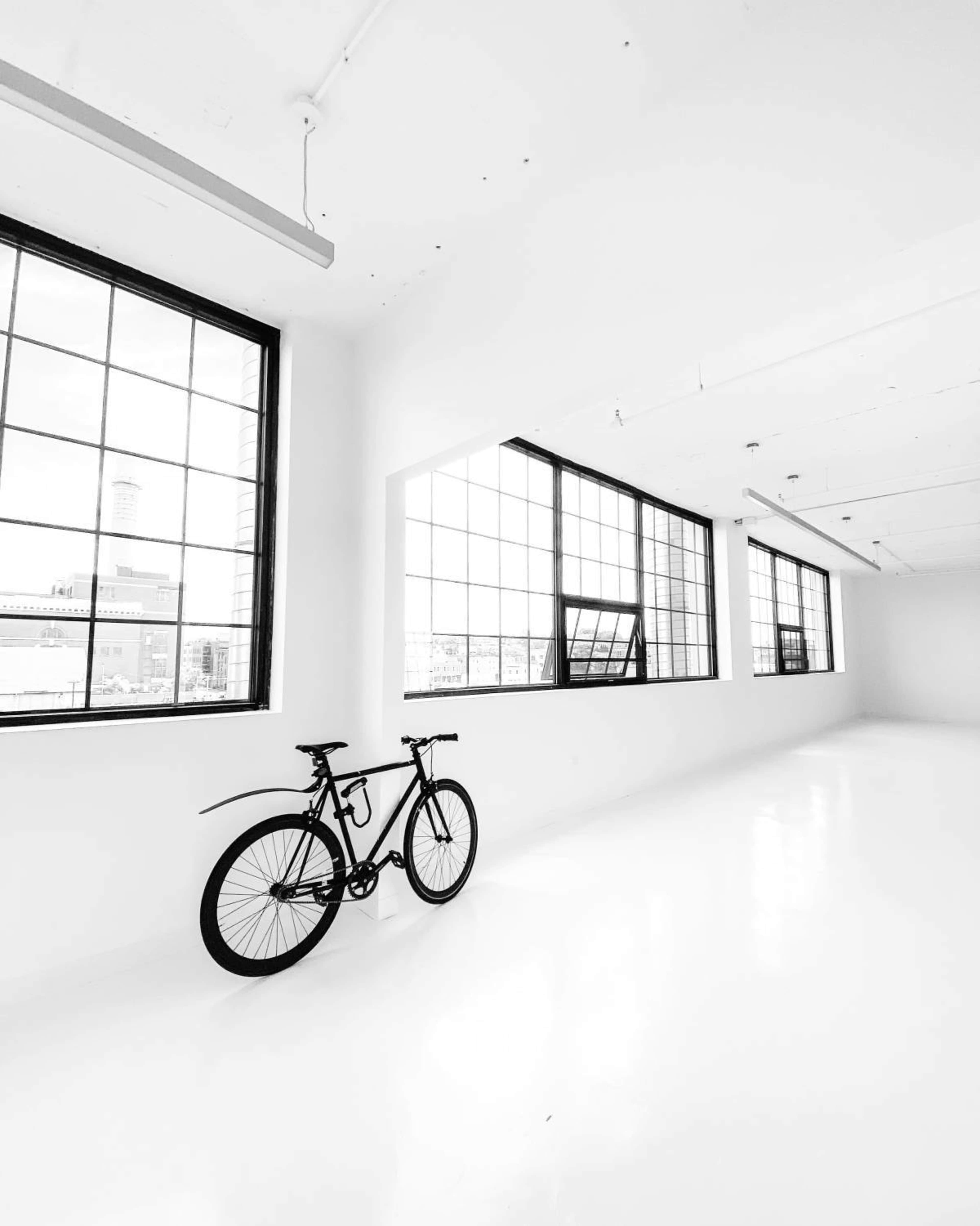 A bicycle is positioned next to a row of large windows in a bright, minimalist room with white walls and a polished floor.