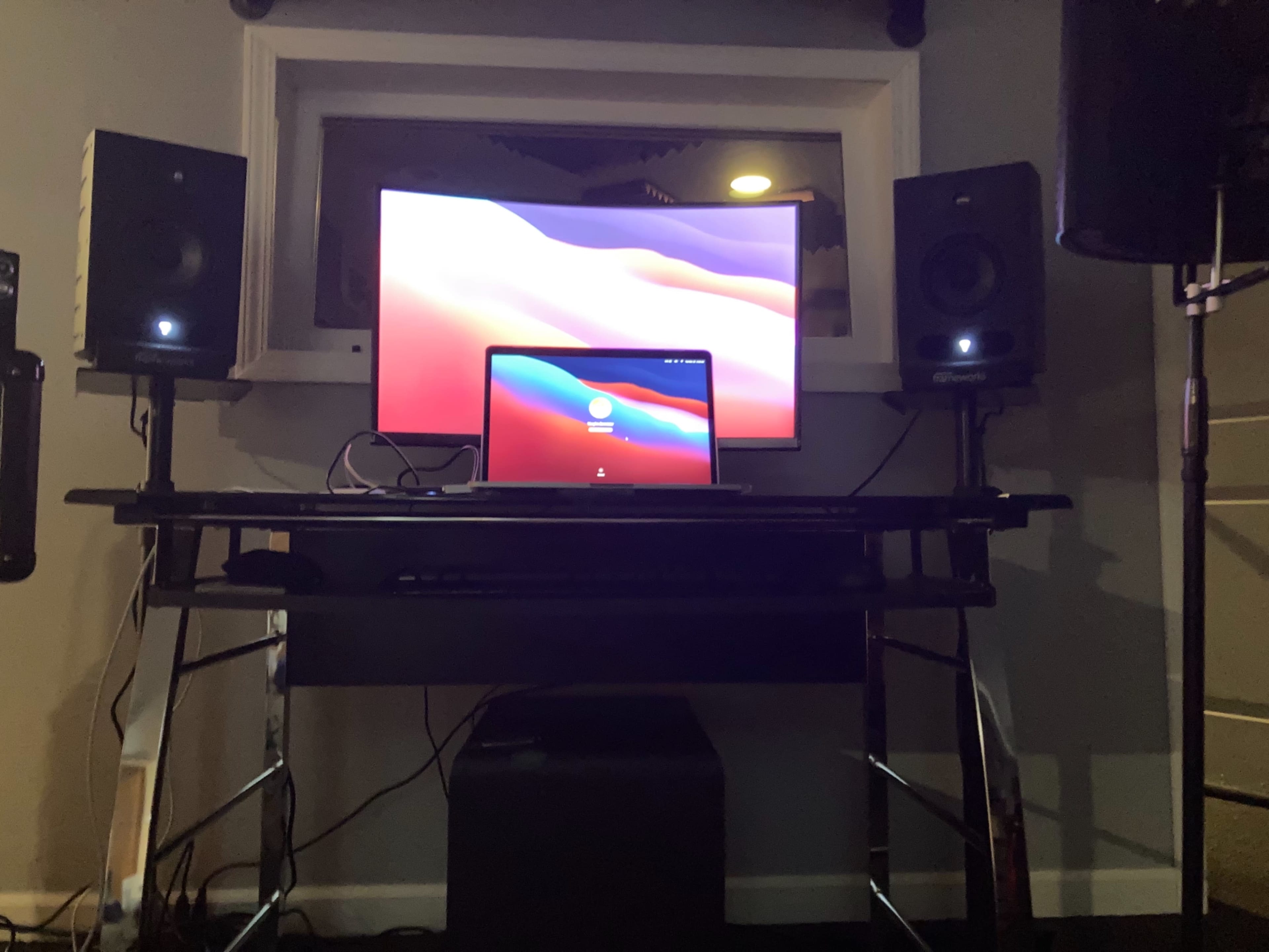 A desk setup with a laptop and a larger monitor, flanked by two speakers, in a room with dim lighting.