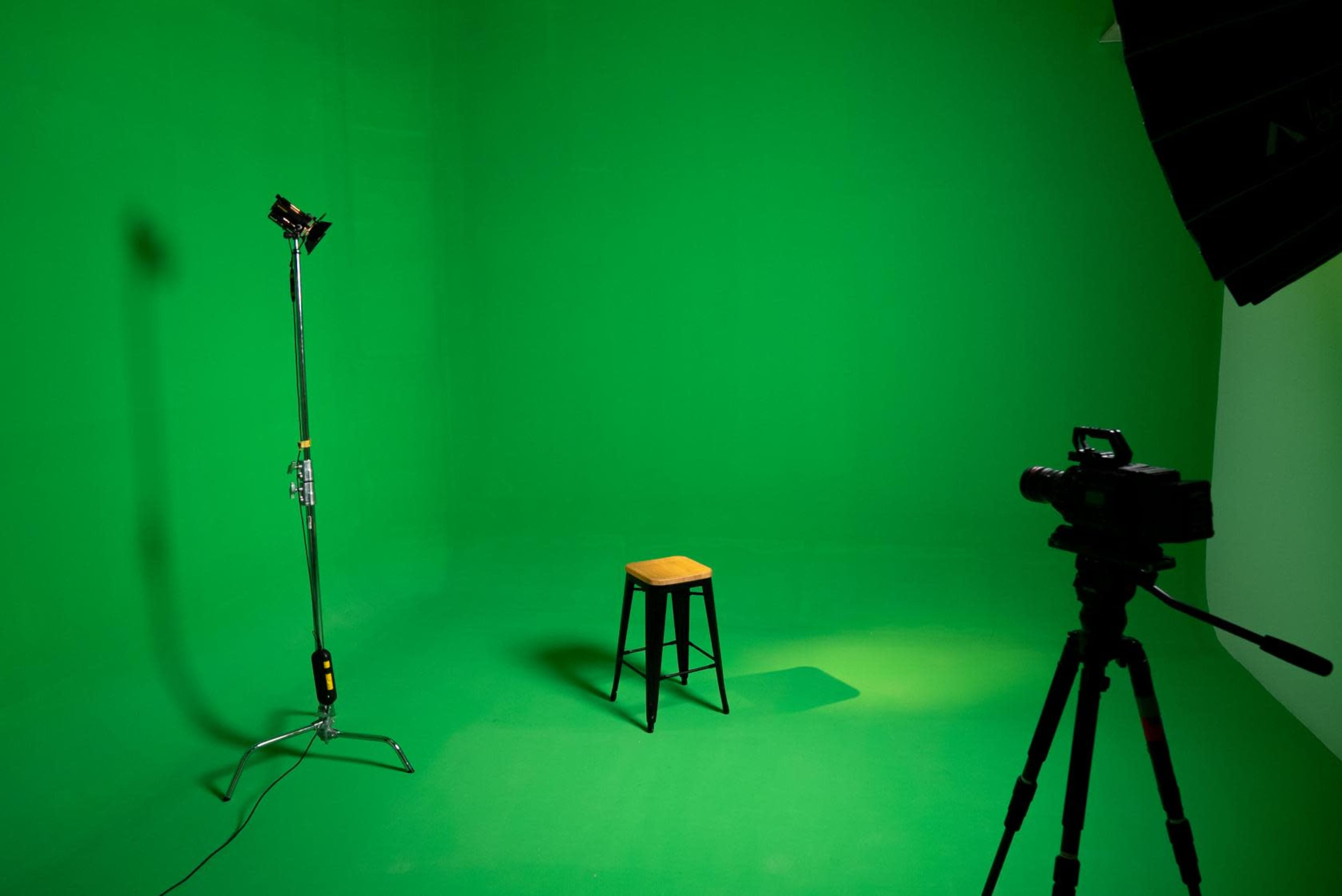 A green screen studio setup featuring a single wooden stool, a light stand, and a camera on a tripod.