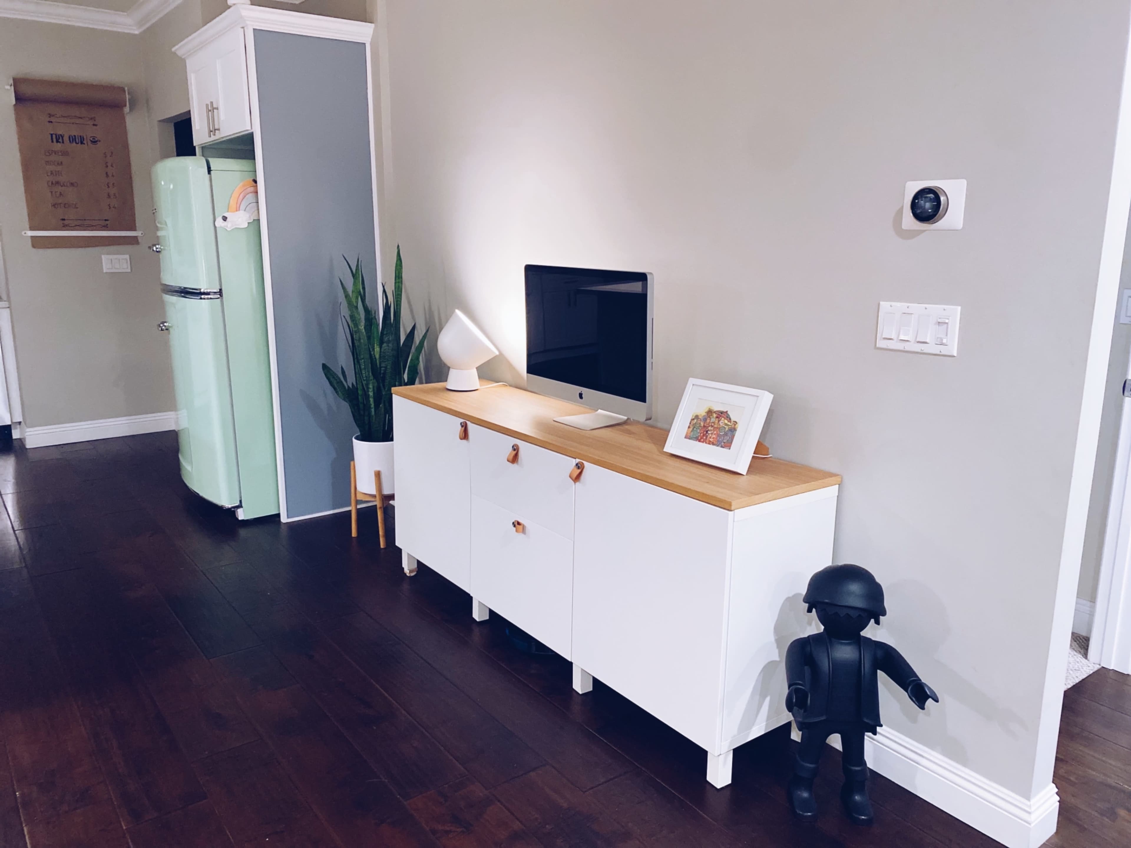 A modern living space features a white cabinet with wooden top, a computer on it, a lamp, and a framed picture, alongside a green refrigerator and a plant in the corner.