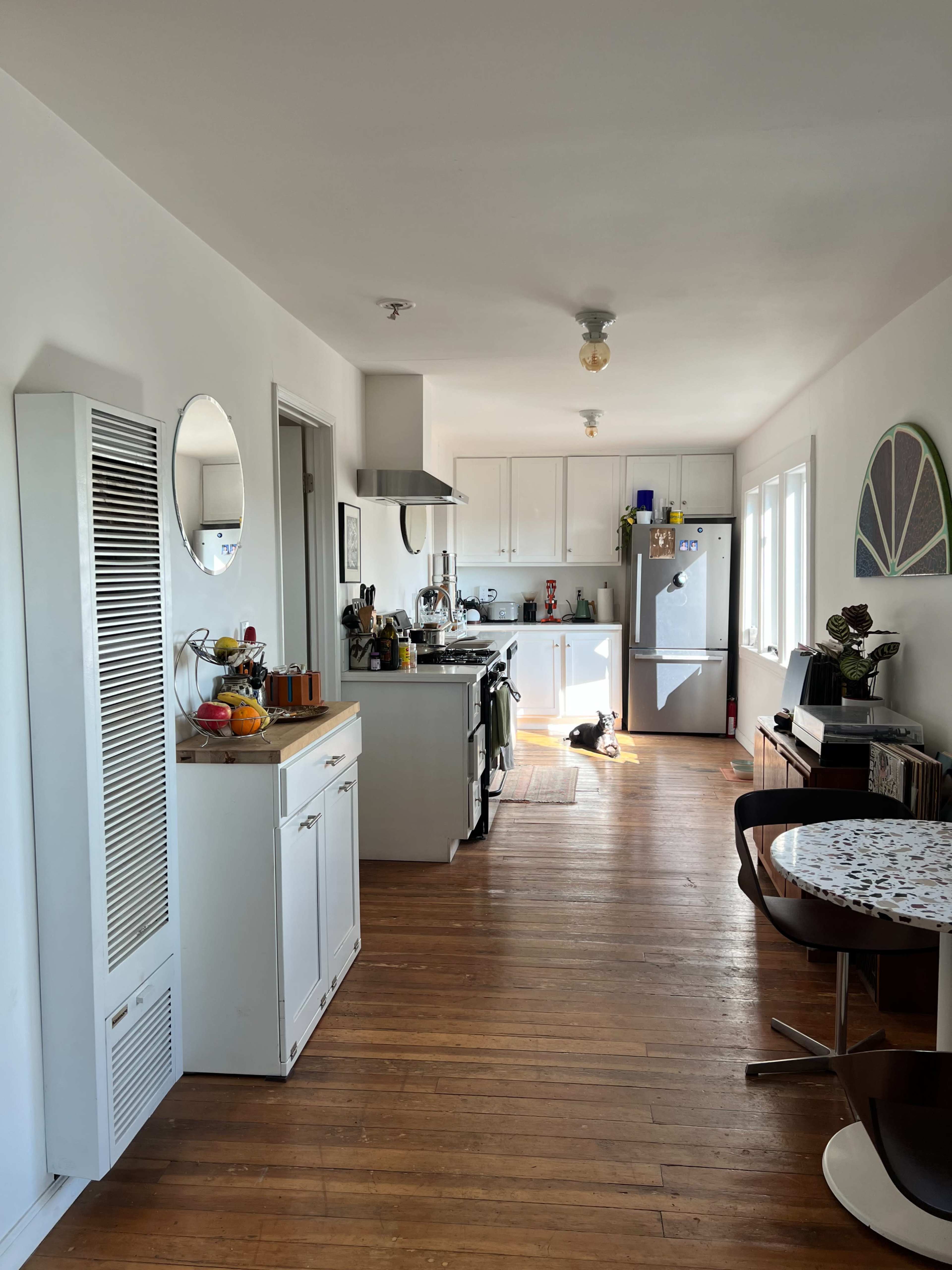 A bright kitchen with wooden floors features a stove, refrigerator, and dining area with a round table.