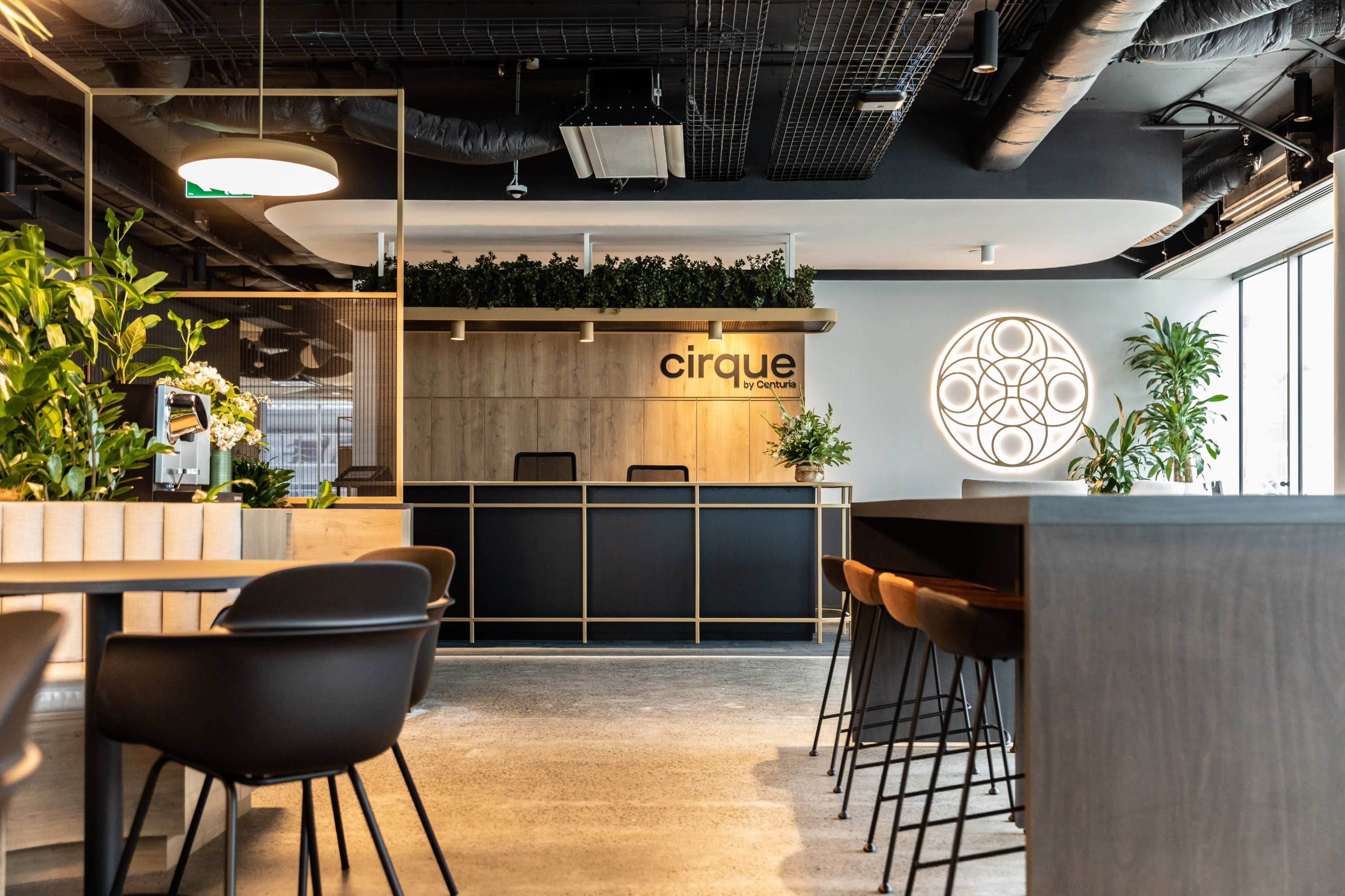 The image shows a modern reception area with a wooden counter, seating, and decorative plants in a well-lit space.
