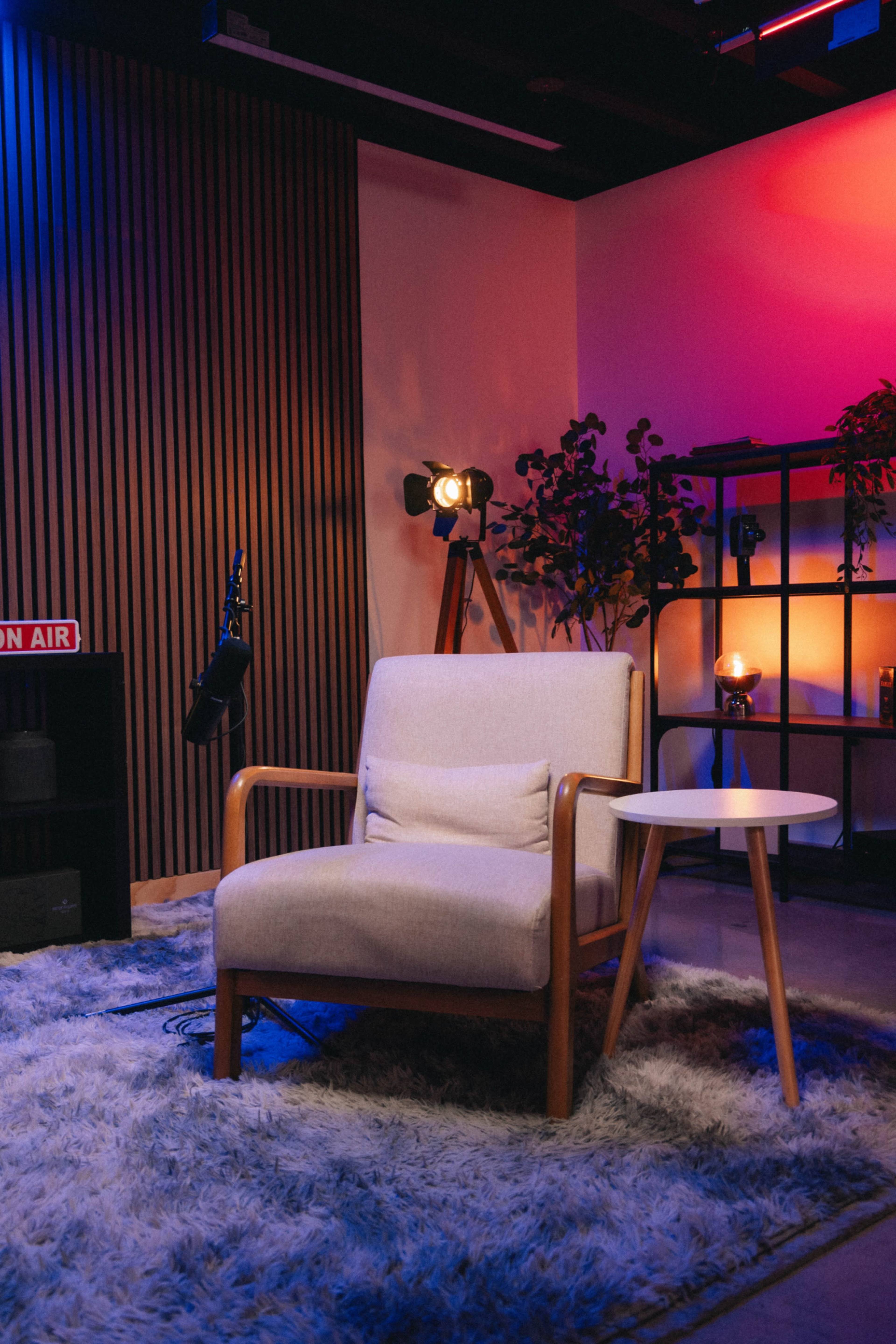 The image features a cozy studio setup with a light-colored armchair next to a small side table, illuminated by colorful lighting and a stylized backdrop.