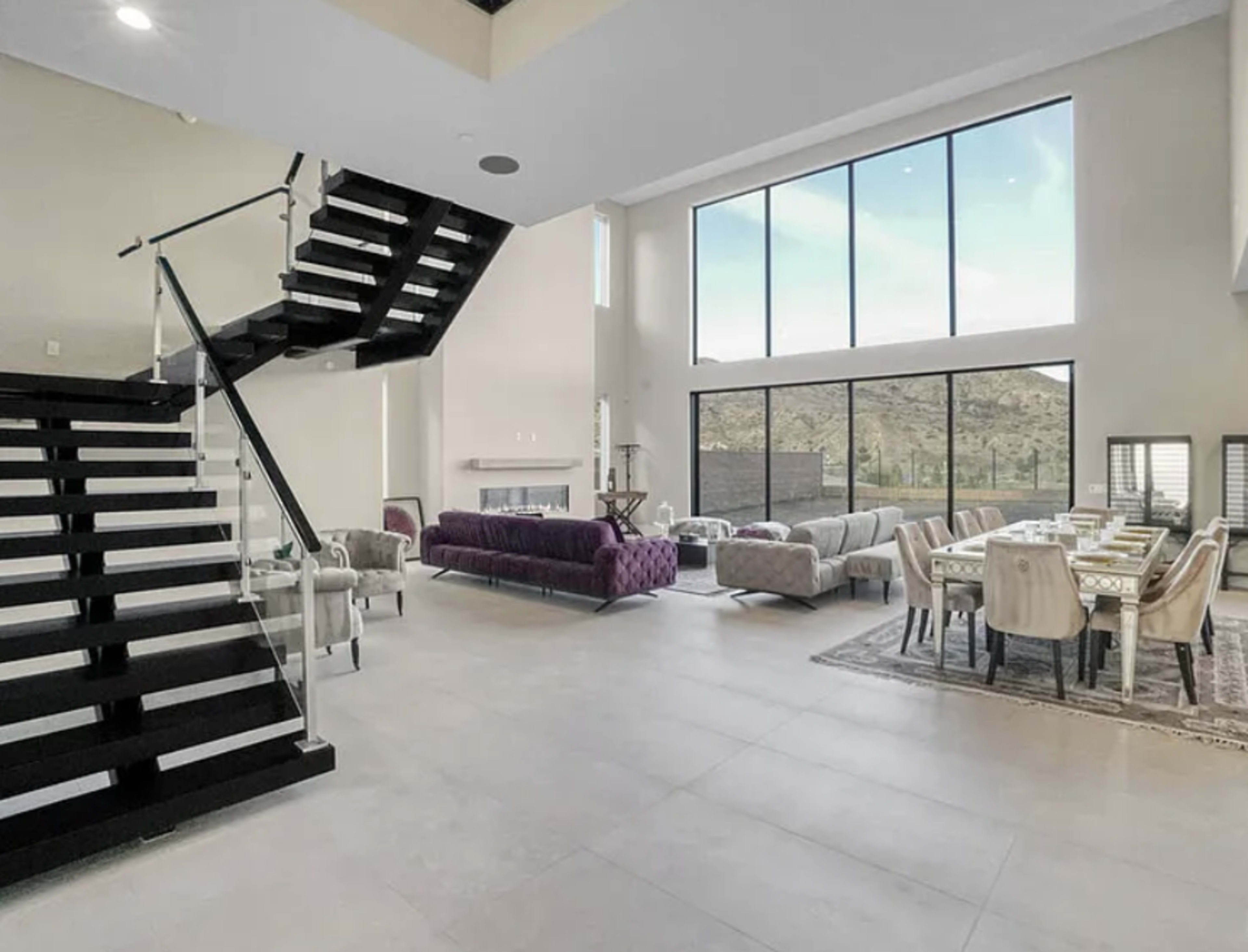 The image shows a modern, open-concept living space featuring a staircase, large windows with a mountain view, and a dining area with a table set for several people.