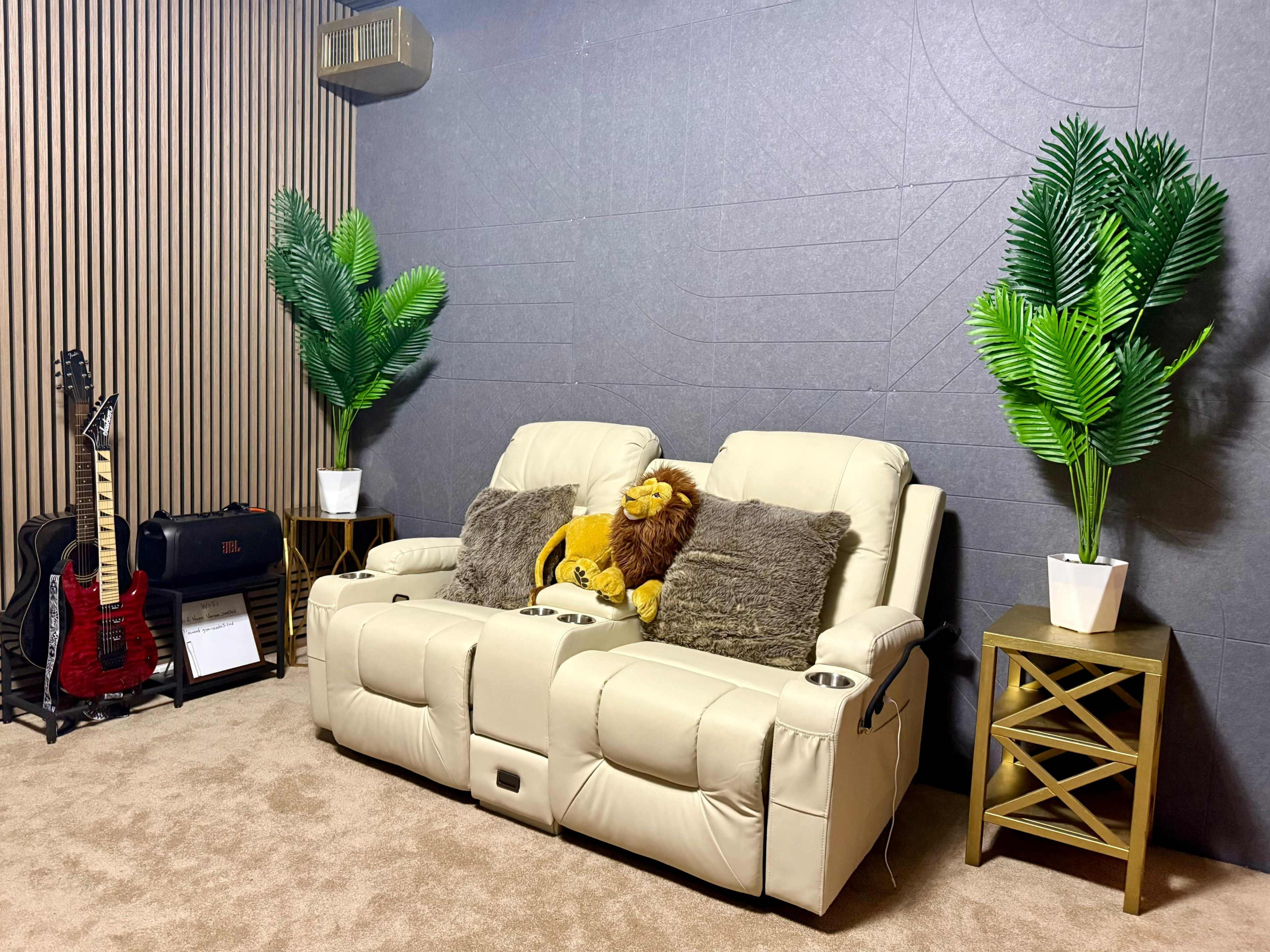 The image shows a cozy living room corner featuring a beige recliner sofa, two potted plants, a guitar stand with an electric guitar, and a decorative side table.