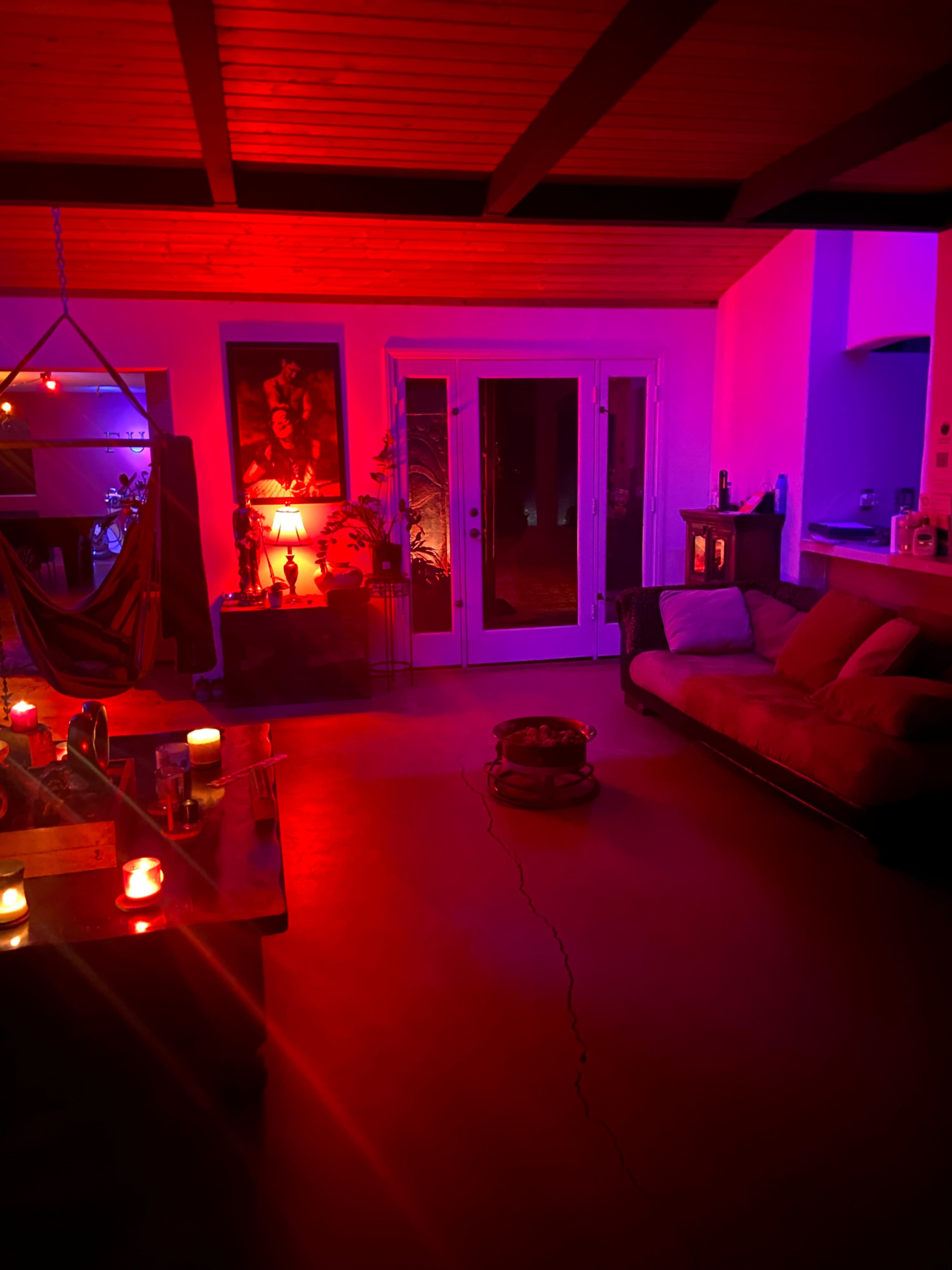 The scene depicts a dimly lit living room with colorful lighting, a couch, a table with candles, and a view of glass doors leading outdoors.