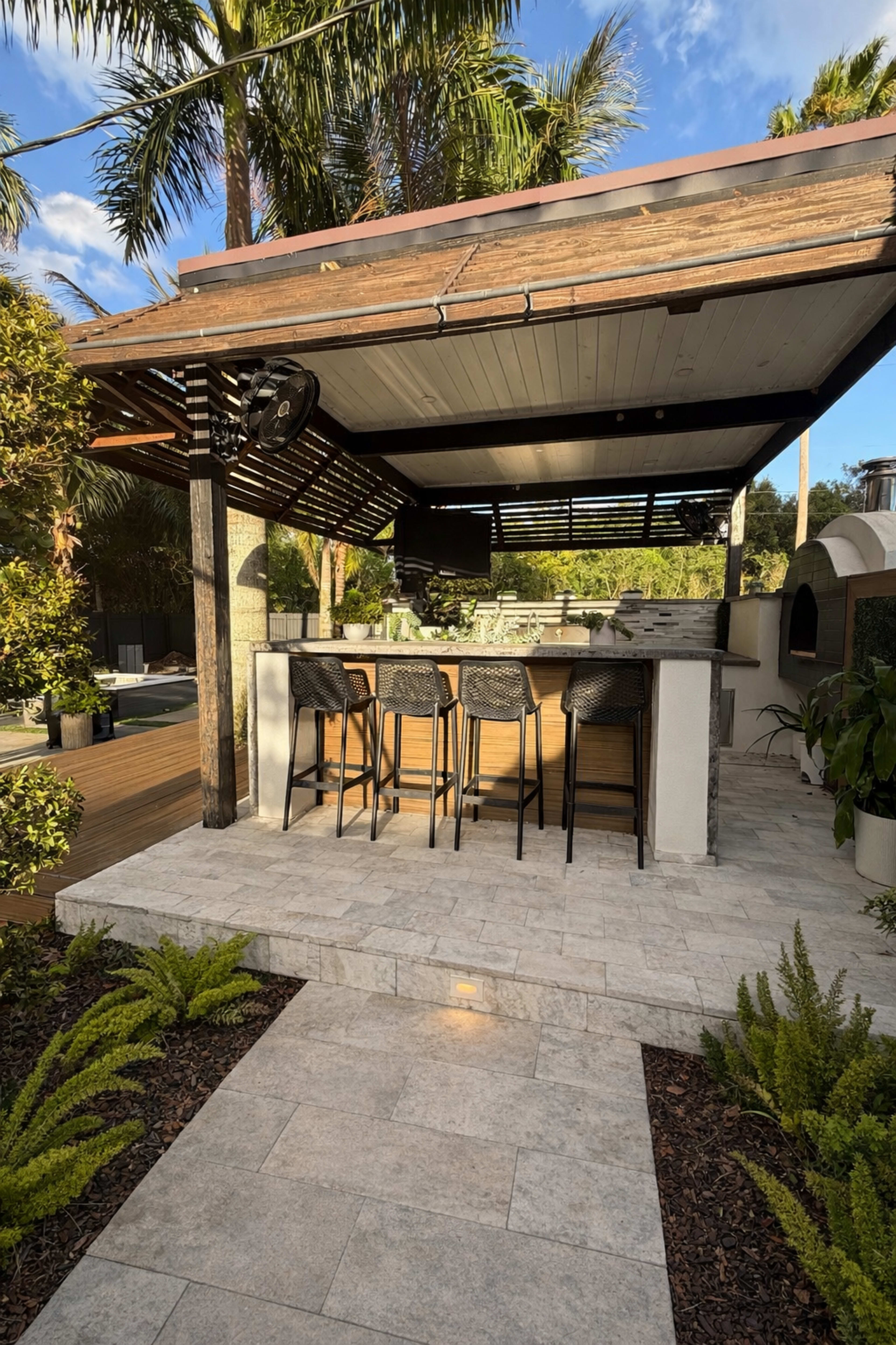 The image shows an outdoor bar area with four bar stools, surrounded by greenery and palm trees, under a wooden pergola.