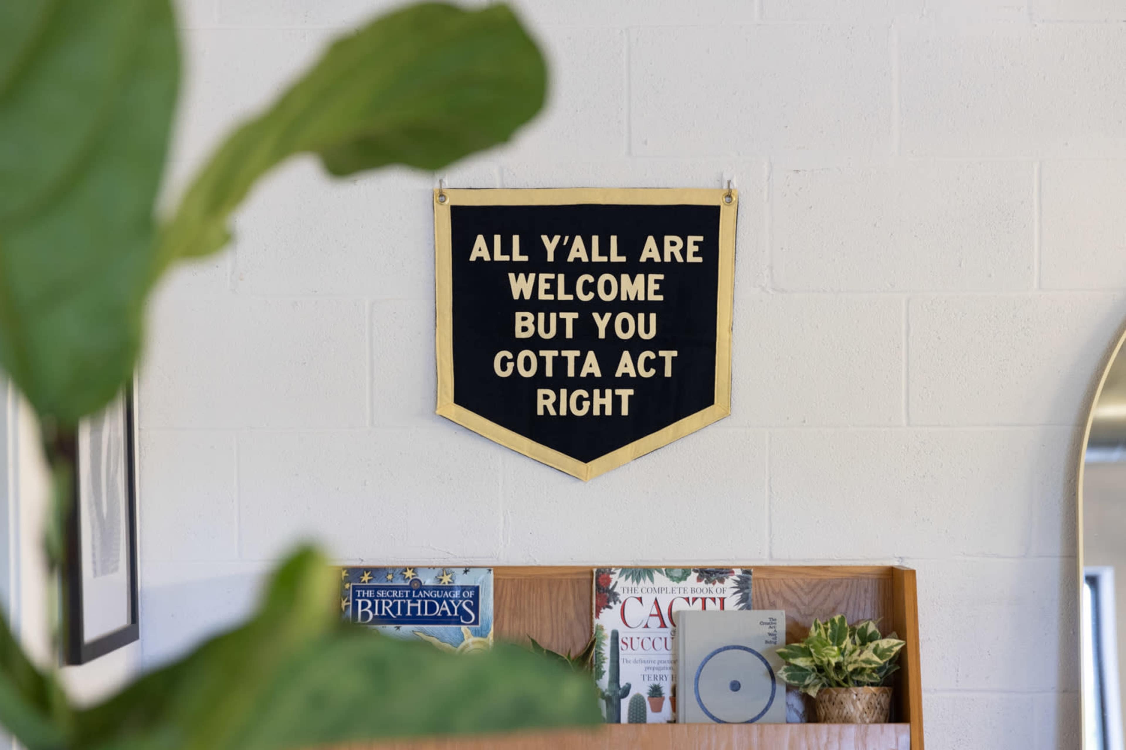 A wall hanging with the text "ALL Y'ALL ARE WELCOME BUT YOU GOTTA ACT RIGHT" is displayed above a wooden shelf containing books and plants.