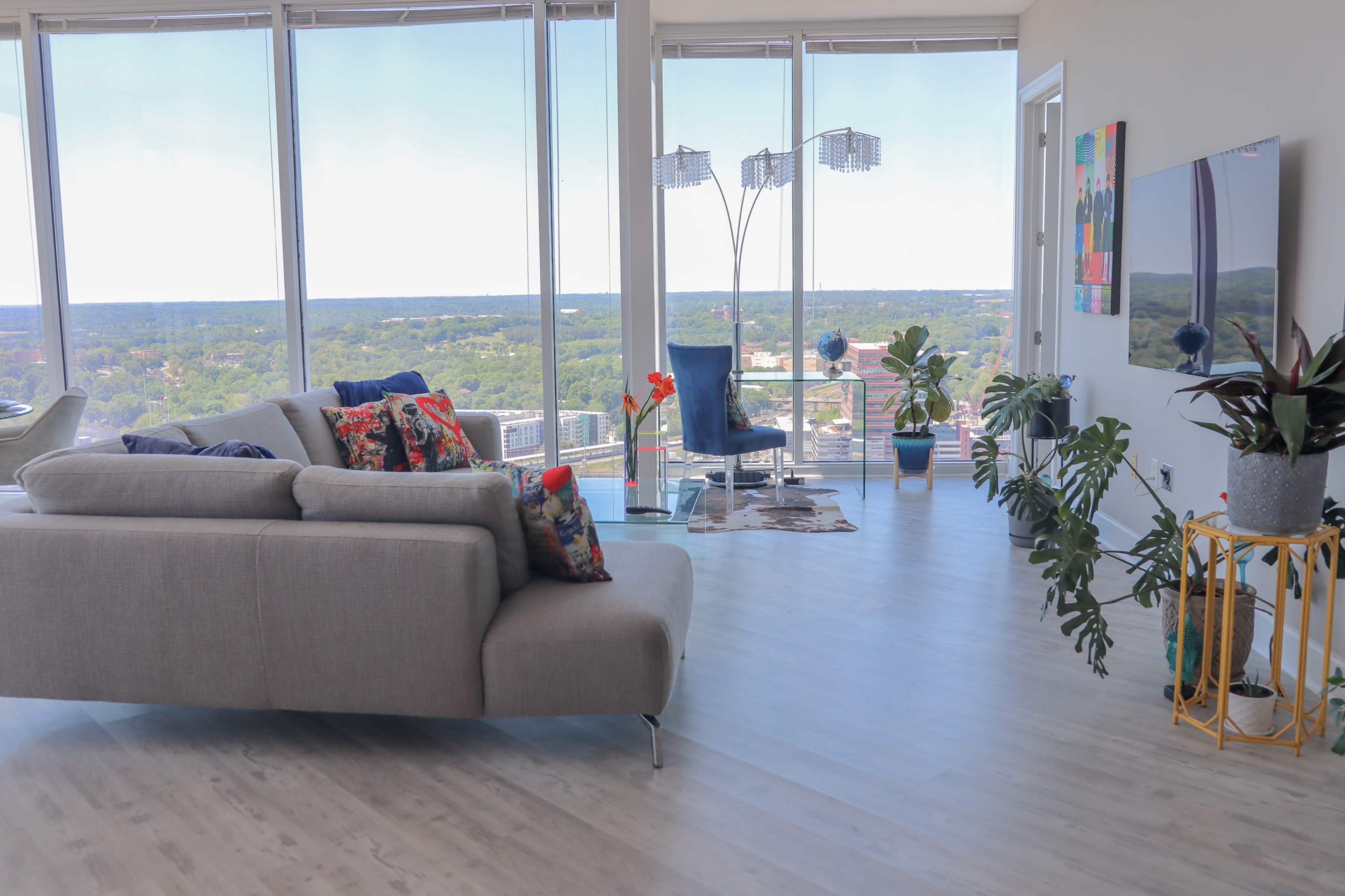 A modern living room features a gray sofa with colorful pillows, glass furniture, and large windows showcasing a panoramic view of the landscape.