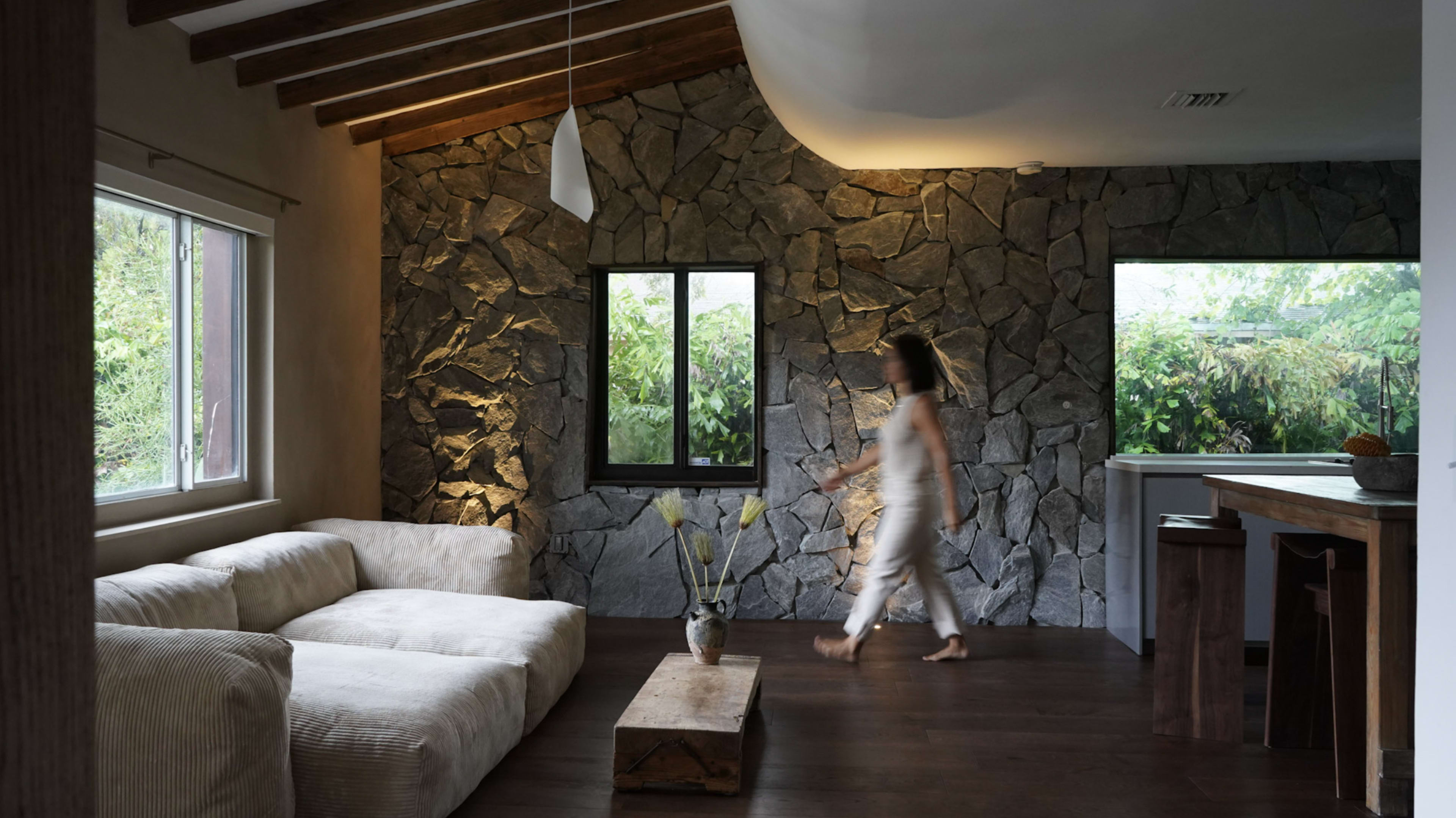 A person walks past a stone wall in a modern living room featuring a light-colored sofa and wooden furniture.