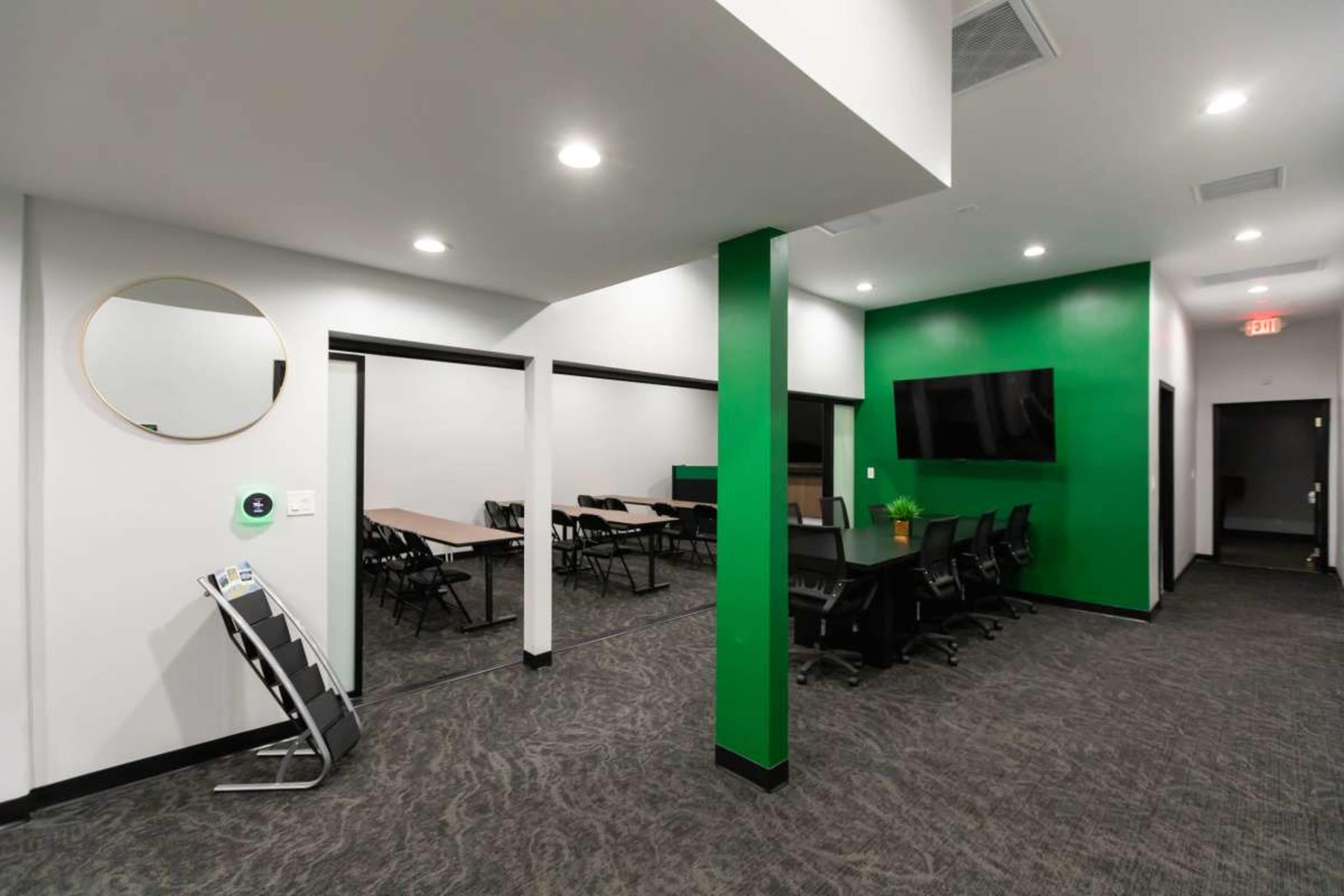 The image shows a modern office space featuring a green accent wall, a large screen on one side, and a meeting area with tables and chairs.