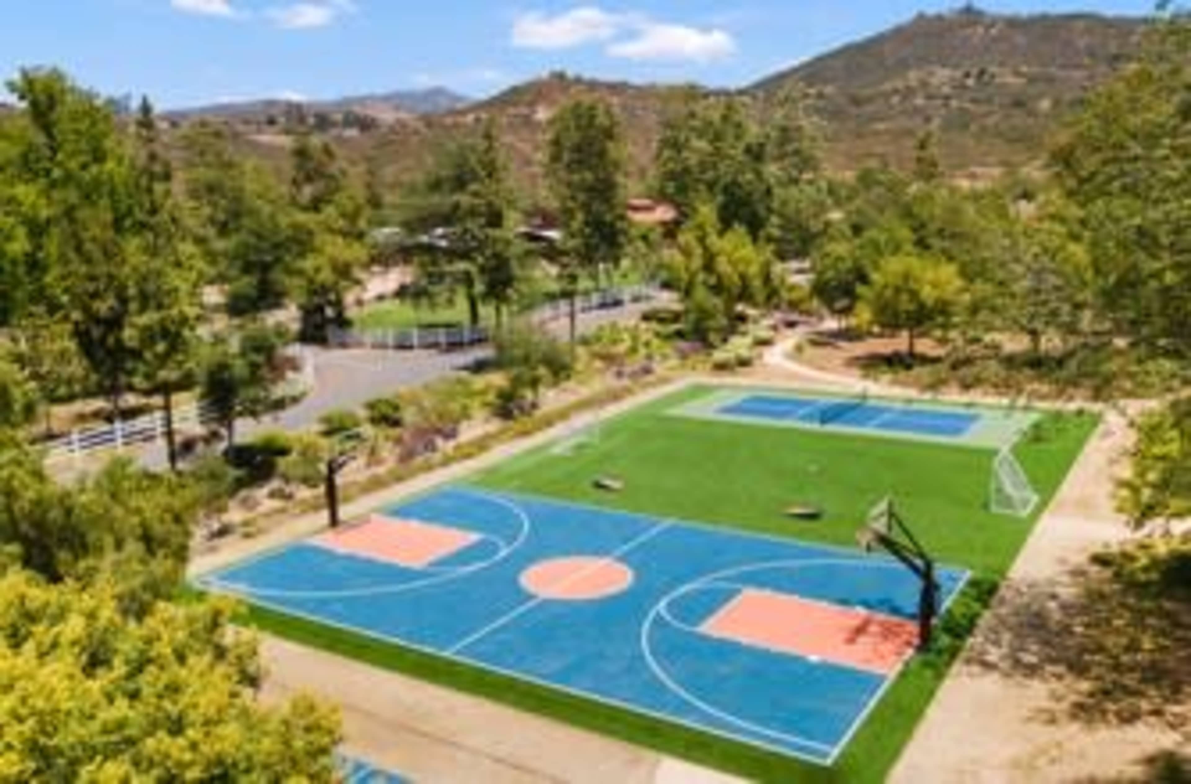 A sports complex featuring two basketball courts and a soccer field, surrounded by trees and hills.