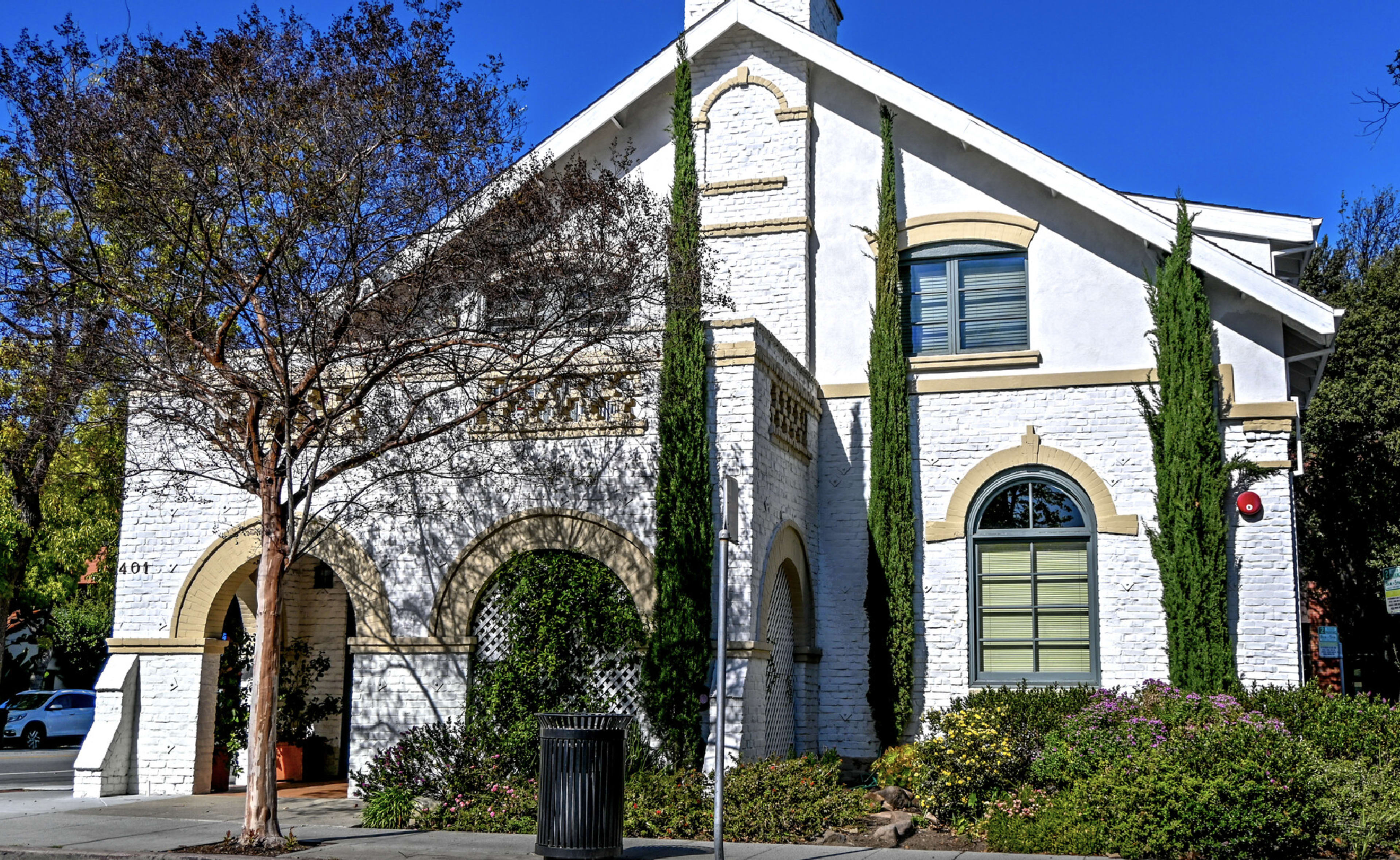 A two-story white building with tall greenery flanking its sides and a decorative facade.