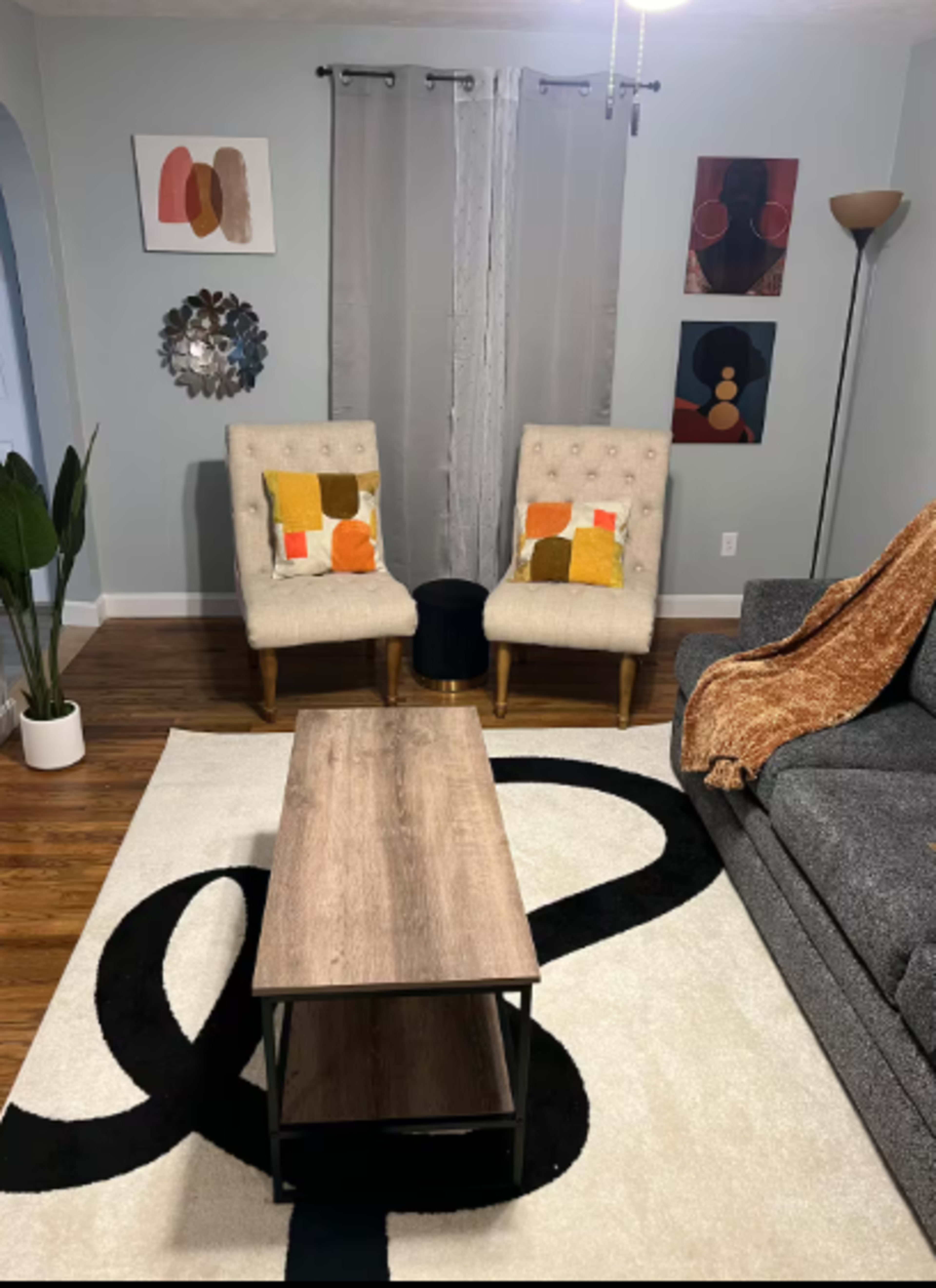 The image shows a modern living room featuring two upholstered chairs, a coffee table, a gray sofa, decorative wall art, and a large area rug.