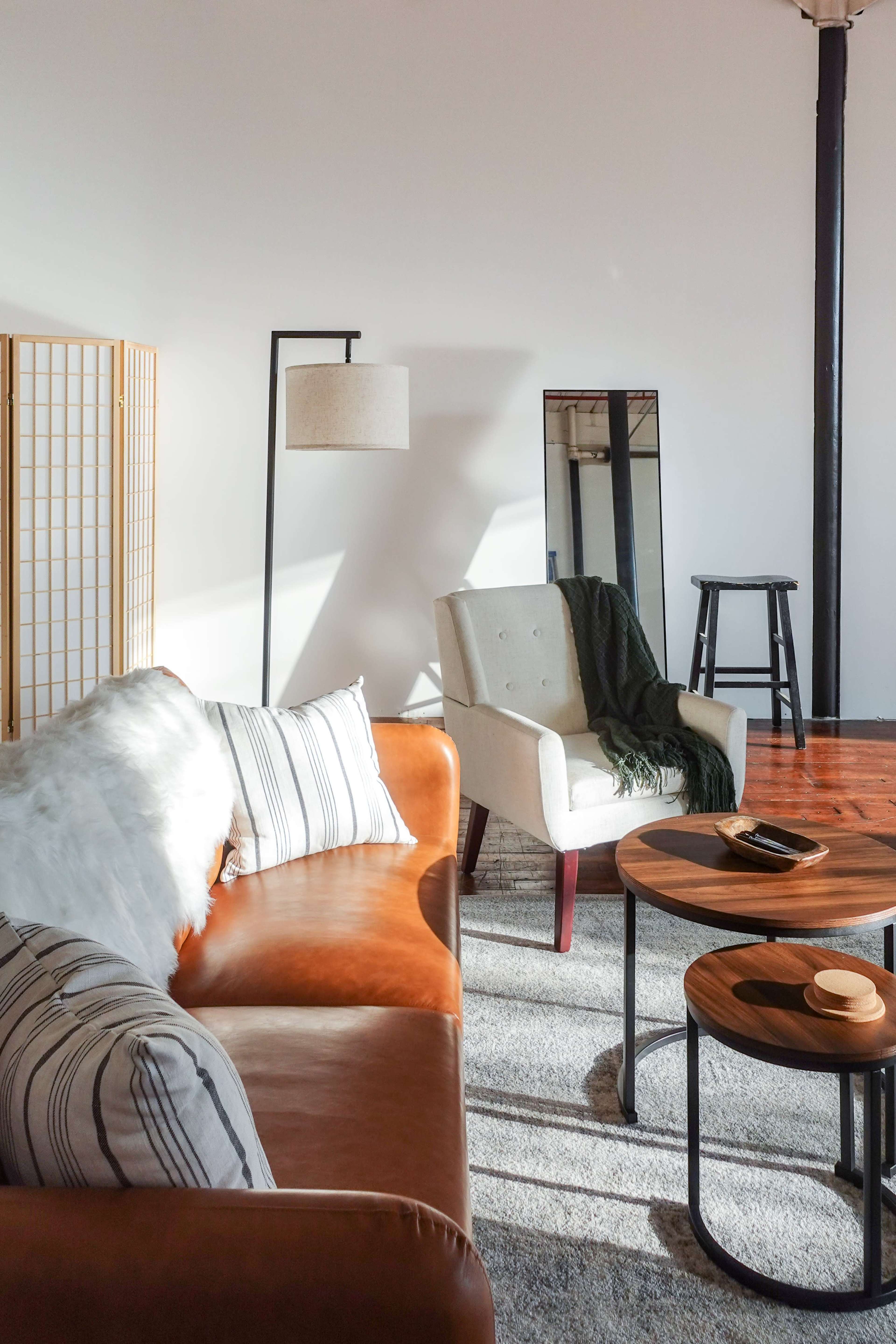 A leather sofa and a light-colored armchair are arranged in a well-lit living room with wooden floors and a large mirror reflecting the space.