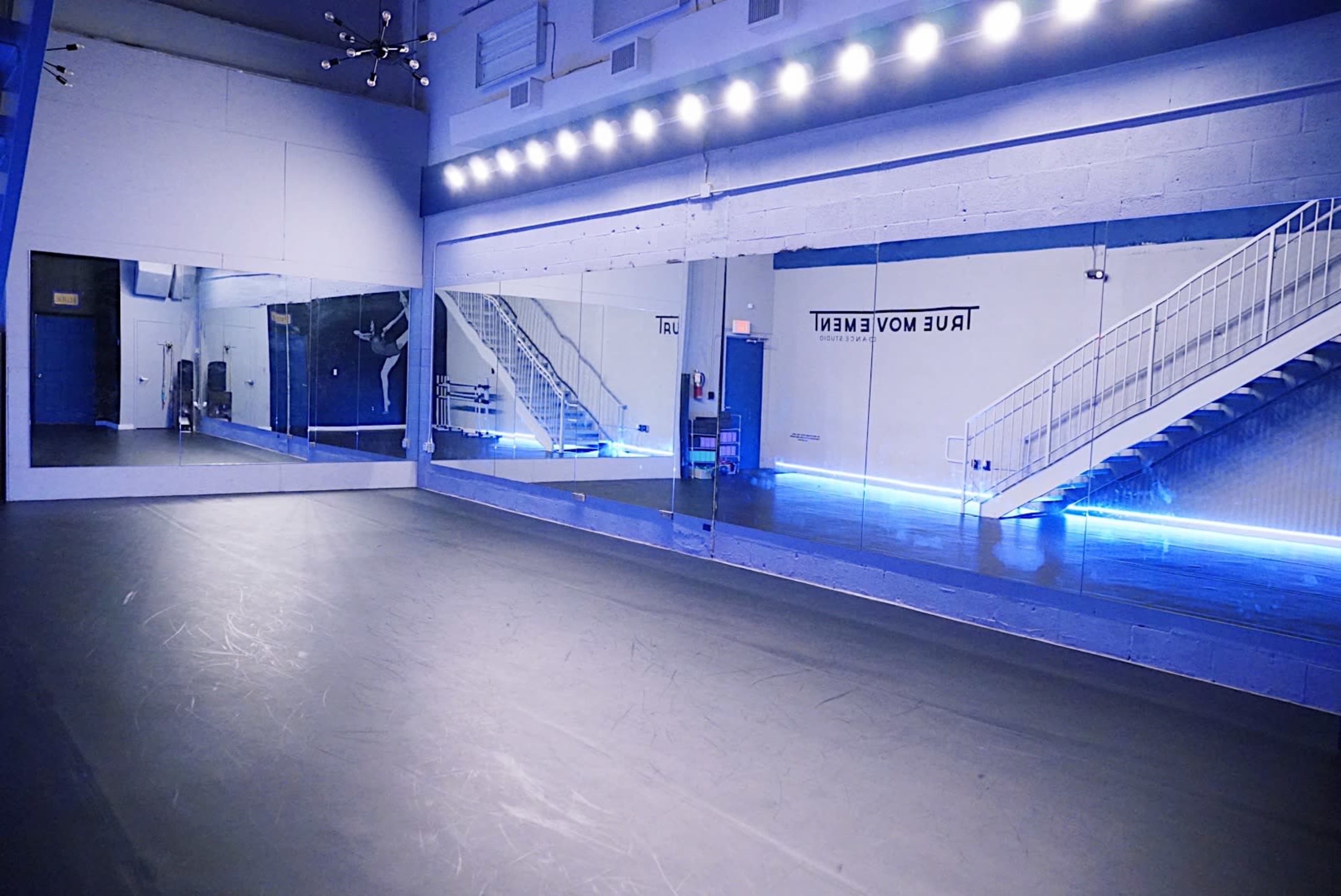 A spacious dance studio features large mirrors, a smooth floor, and soft blue lighting.