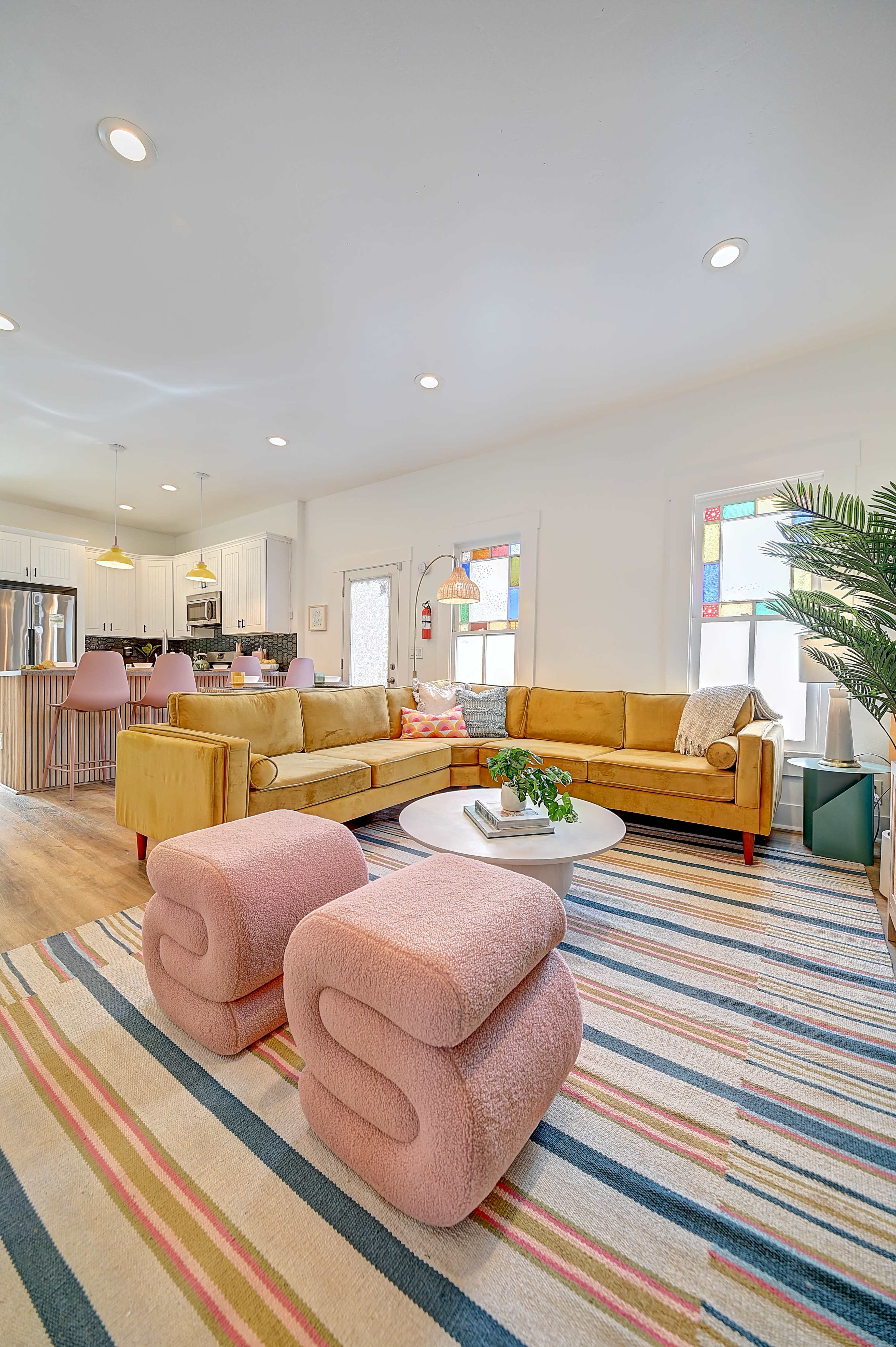 The image shows a modern living room featuring a yellow sectional sofa, a round coffee table, and pastel-colored stools on a striped area rug.
