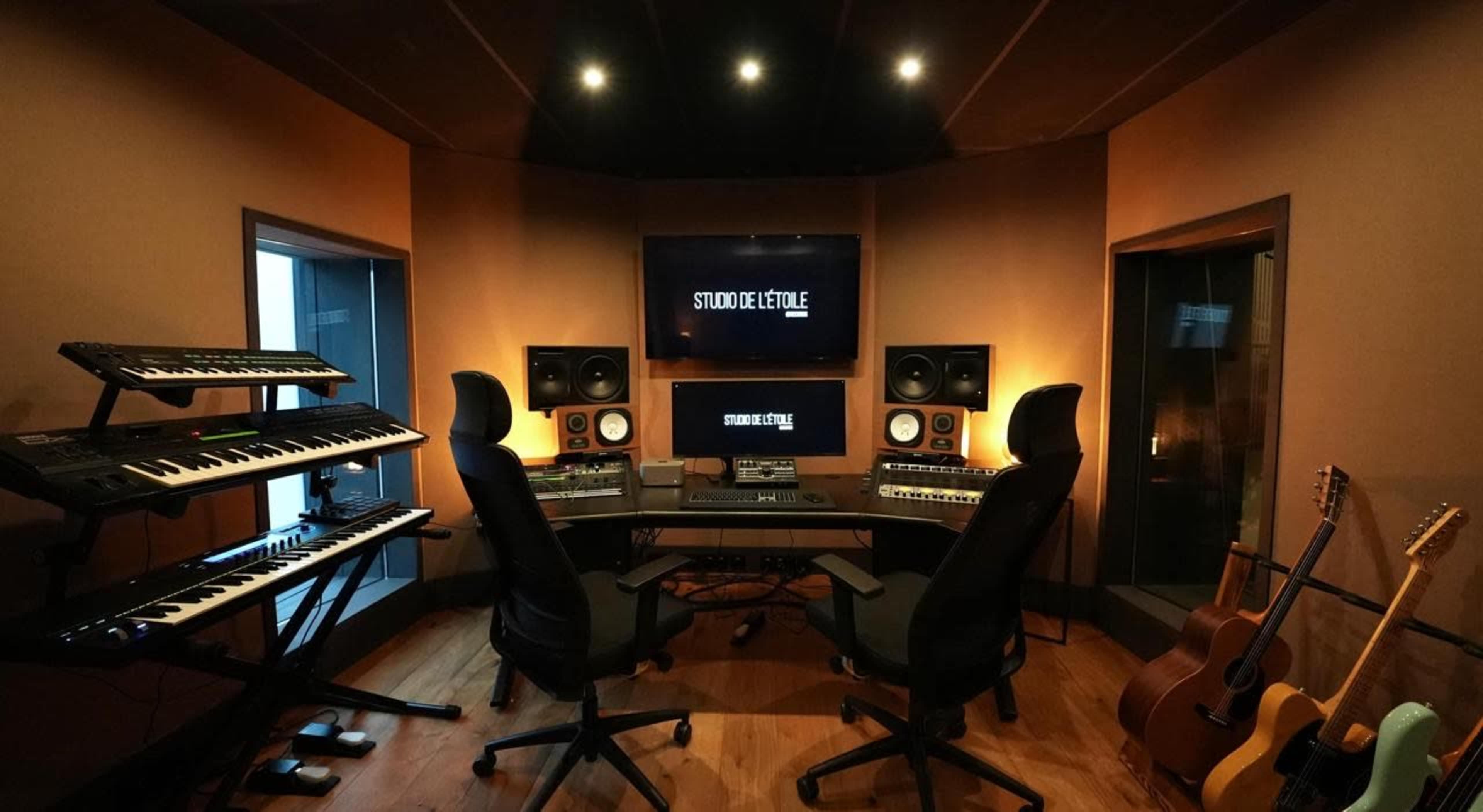 A music production studio features two black swivel chairs, a control desk with various audio equipment, two speaker monitors, and keyboards, all set in a warmly lit room with acoustic panels.