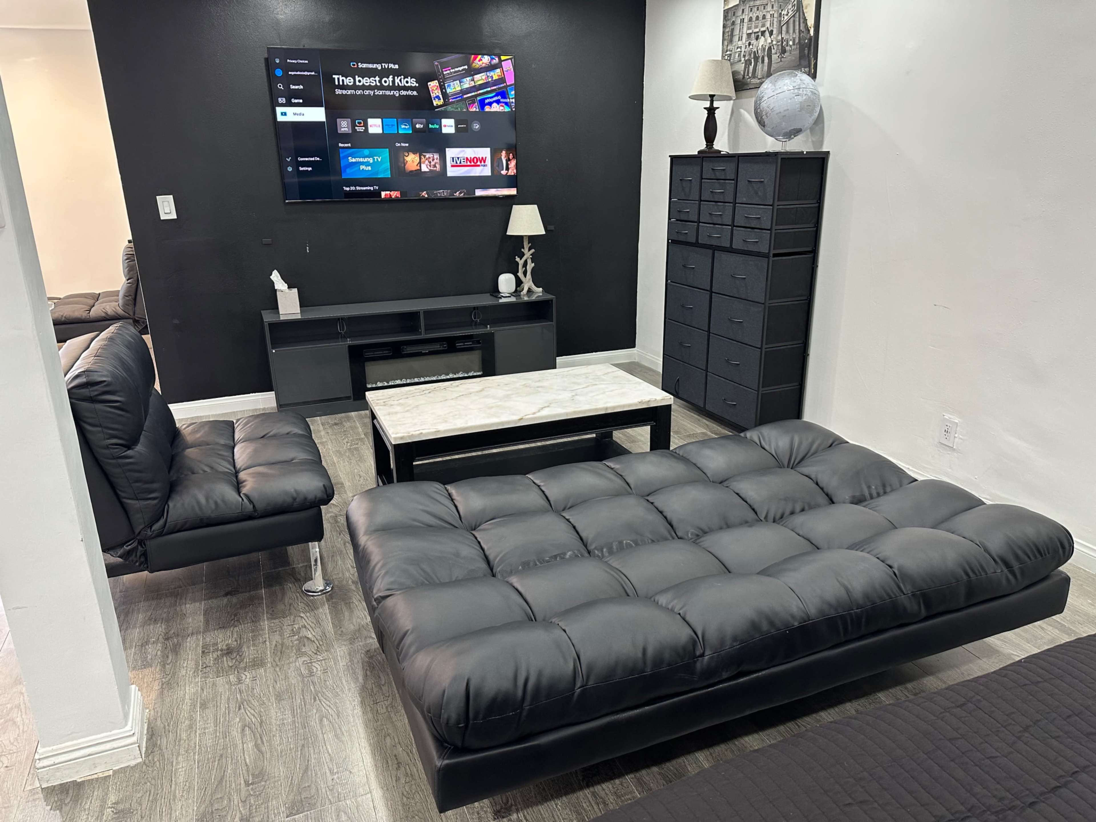 The image shows a modern living room with a black sofa, a marble-topped coffee table, a TV mounted on the wall, and a tall dresser in the background.