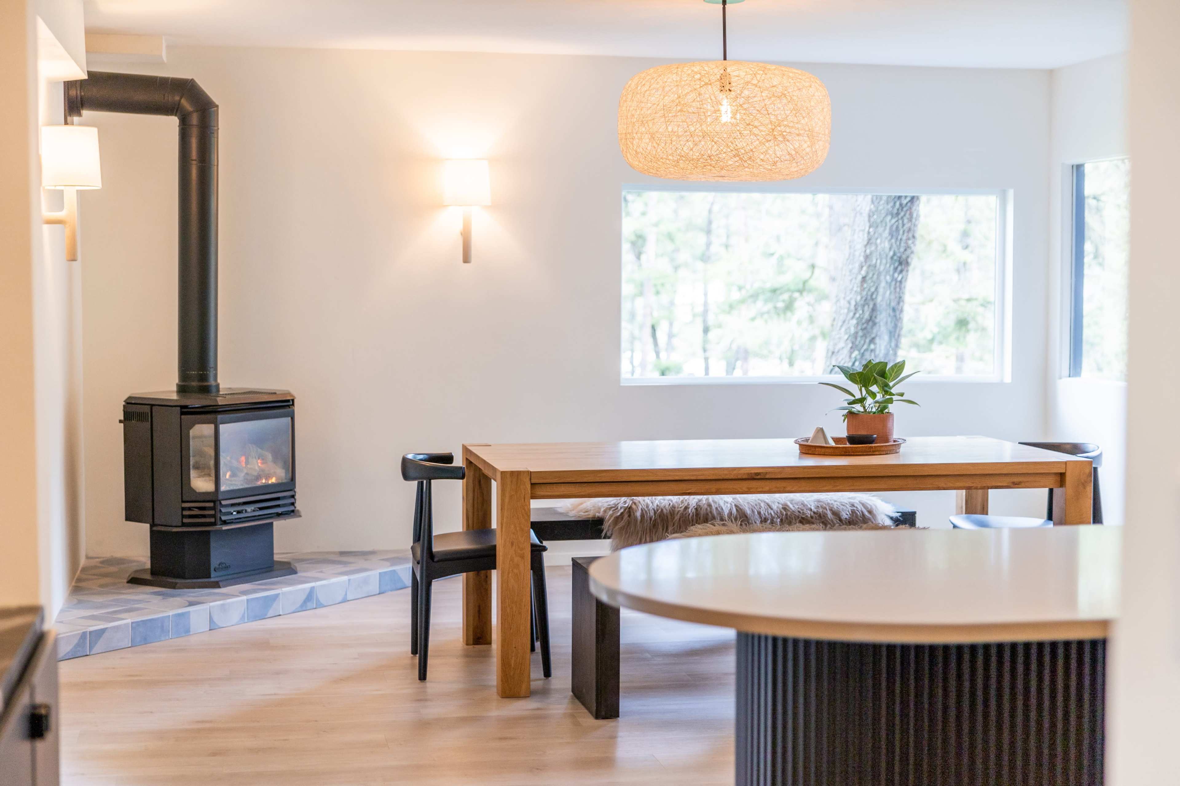 The image shows a modern dining area with a wooden table, black chairs, and a wood-burning stove, surrounded by large windows that allow natural light to enter.