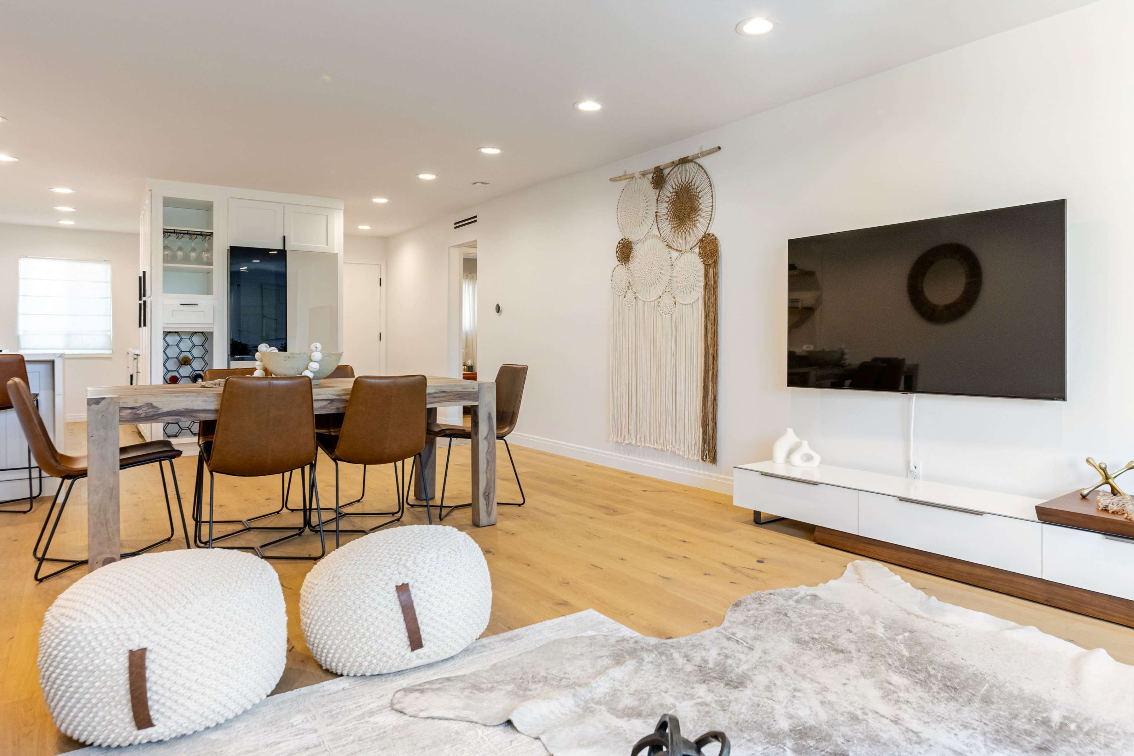 A modern living space features a dining area with a wooden table and brown chairs, a television mounted on the wall, and decorative elements including a macramé wall hanging and round poufs.