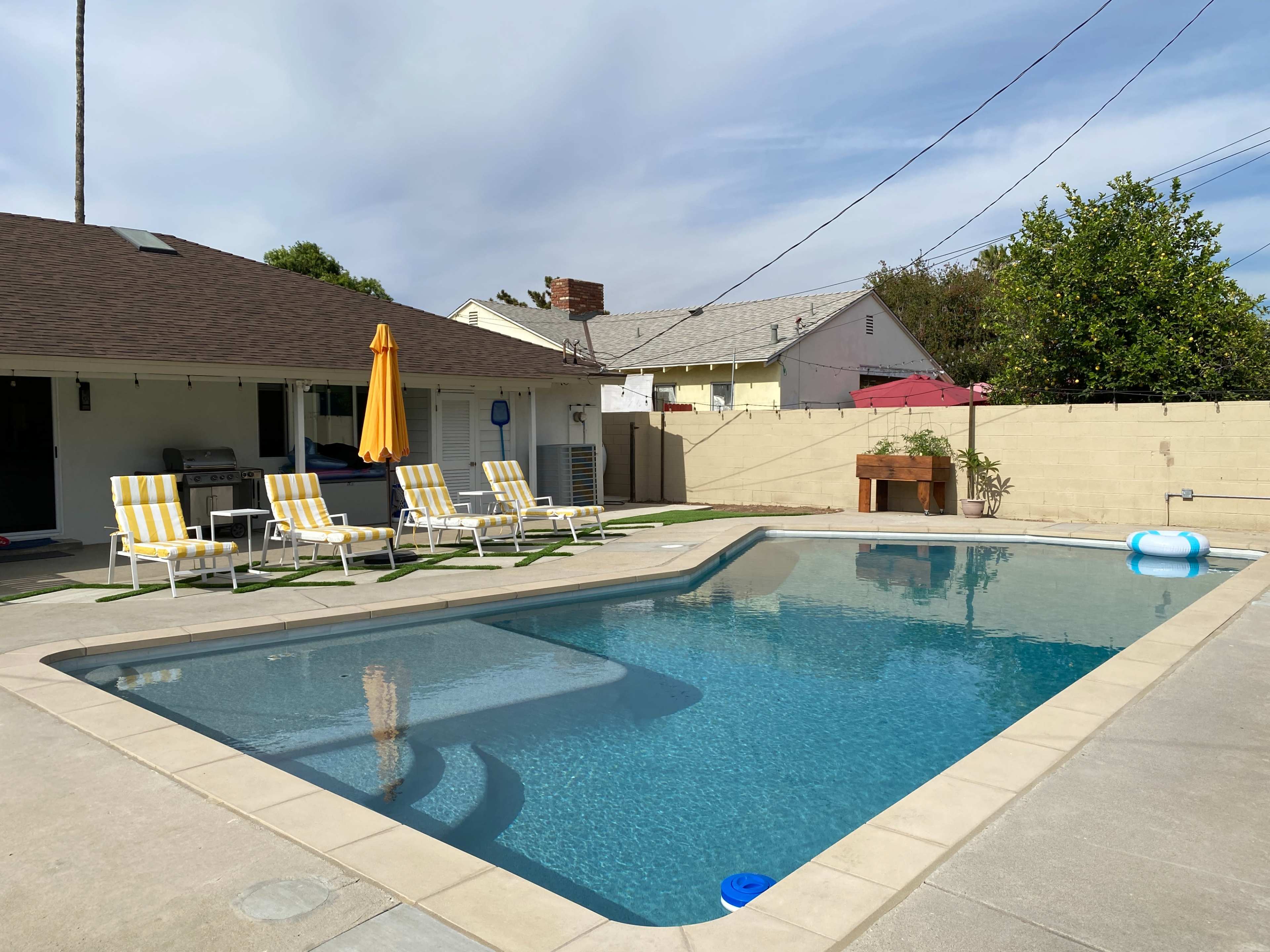 A backyard swimming pool surrounded by lounge chairs, with a yellow umbrella and a barbecue area in a residential setting.