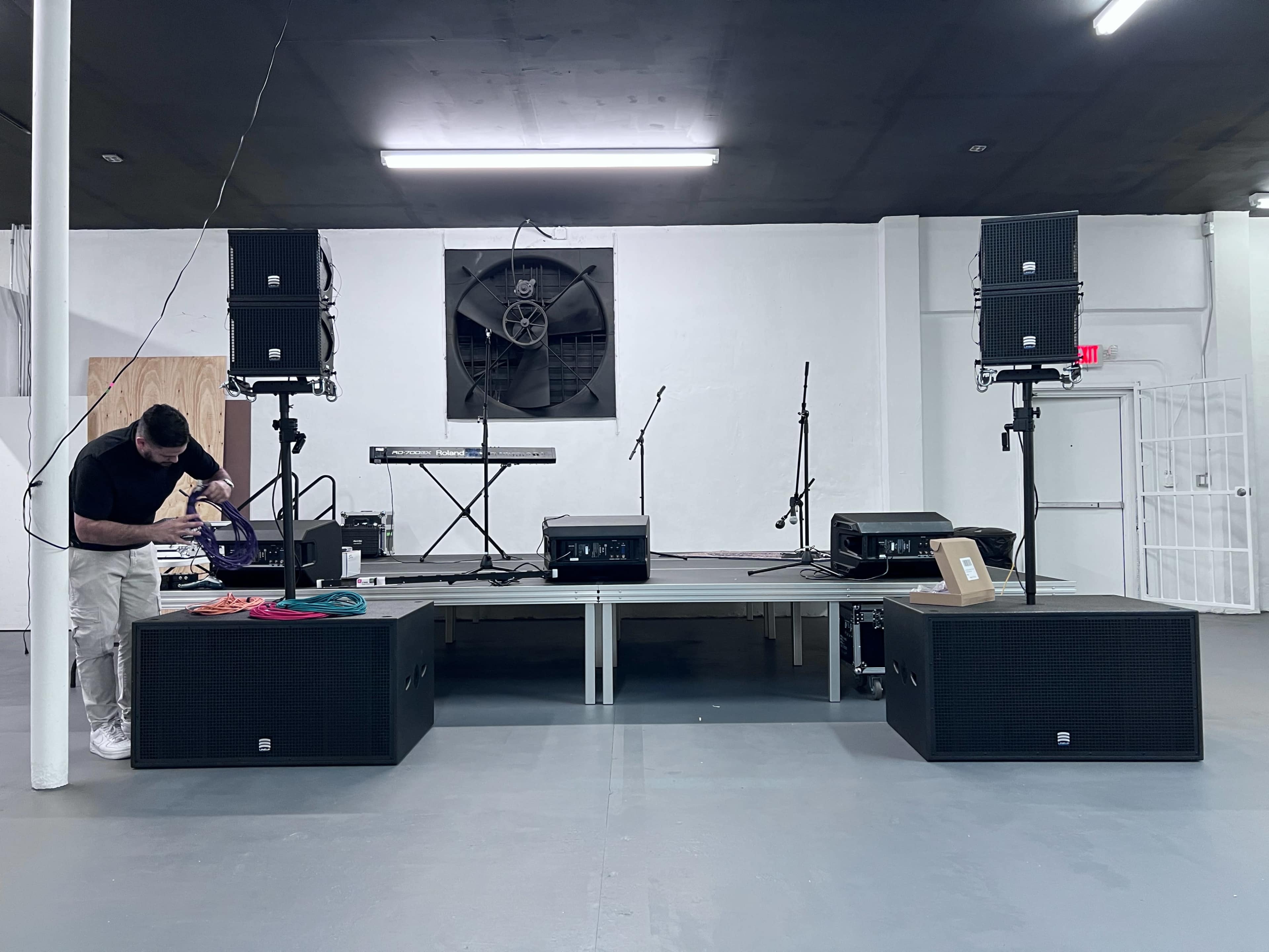 A stage setup is shown with two large speakers on either side, a keyboard on a stand in the center, and microphones positioned for an upcoming performance.