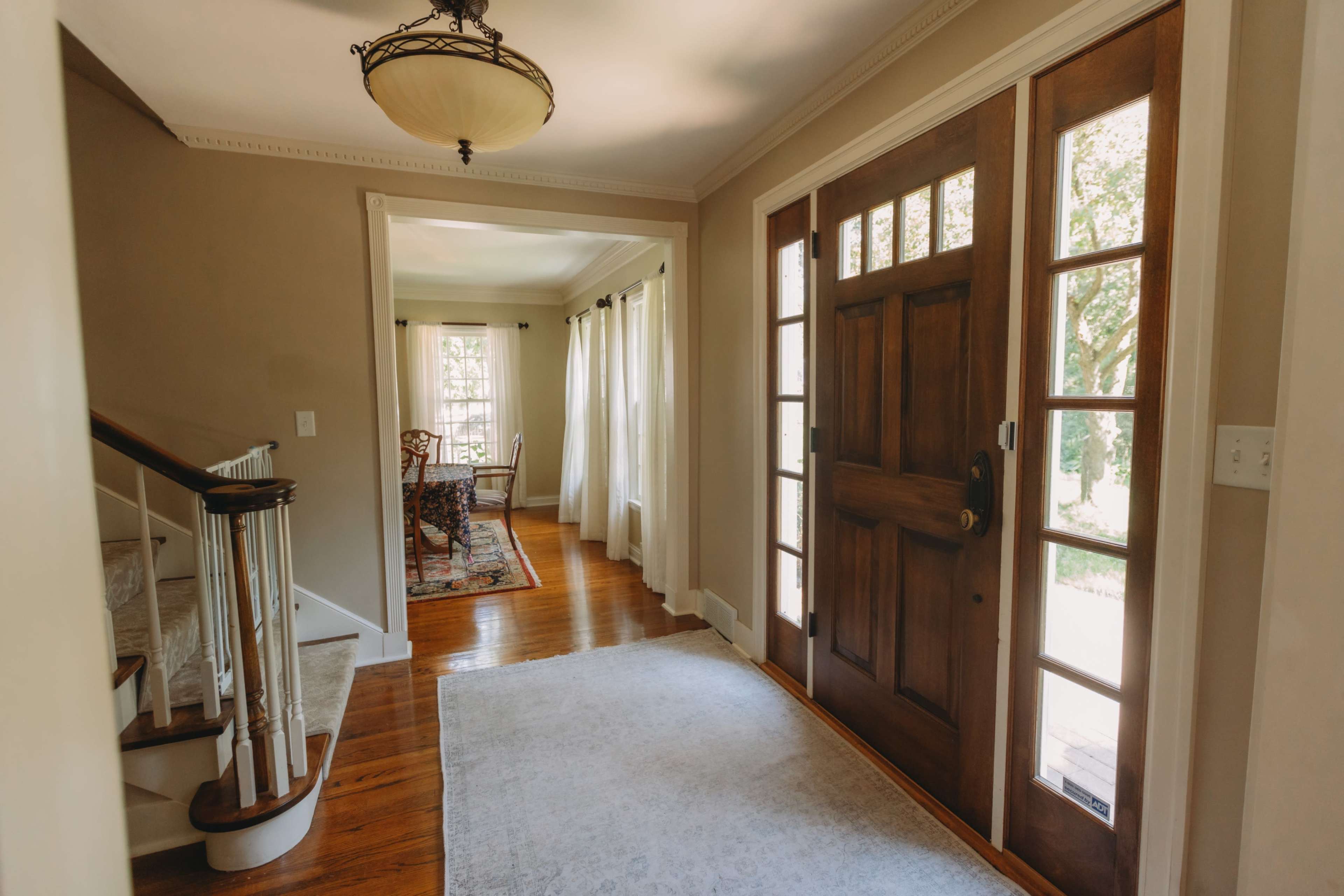 The image shows a bright entryway featuring a wooden front door, a staircase, and a view into a dining area with large windows.