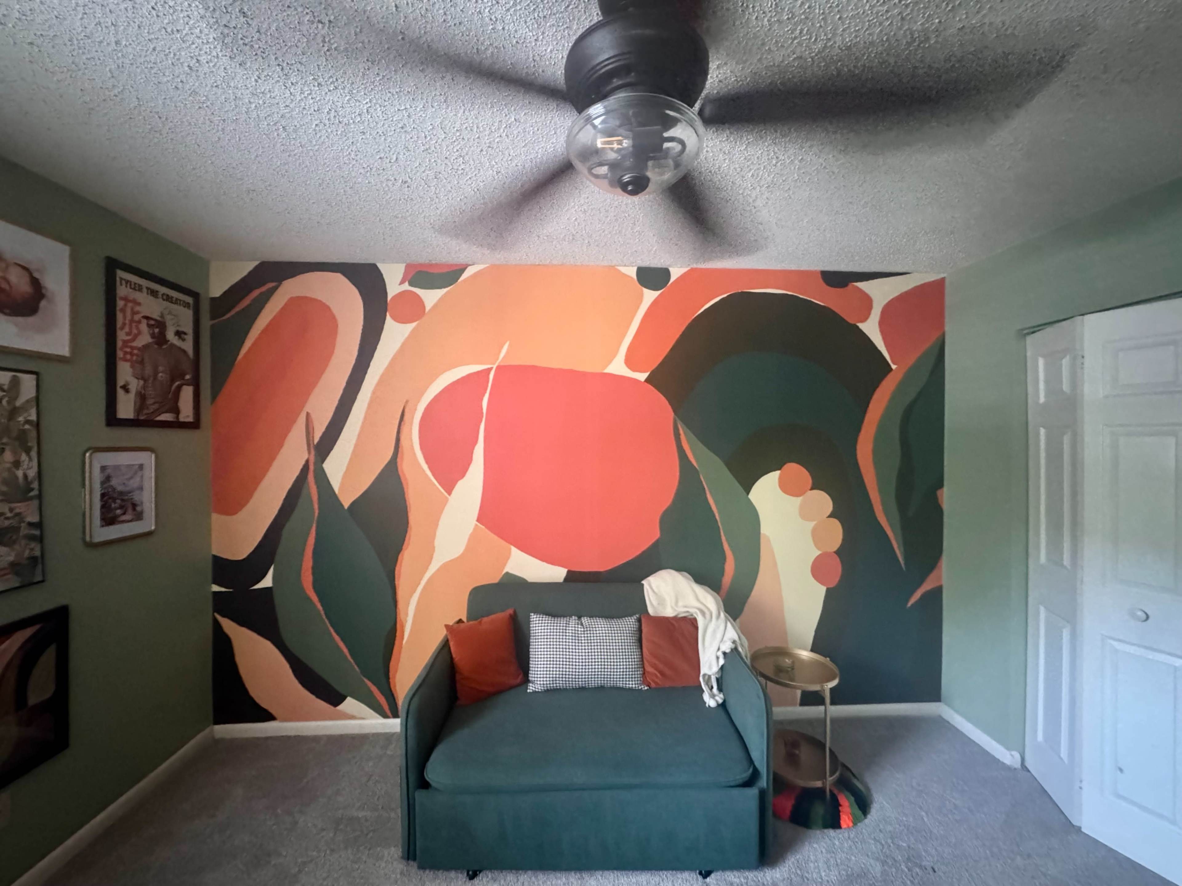 The image shows a room with a green wall, a large abstract mural on one wall, a green couch with colorful pillows, a small table, and a ceiling fan.
