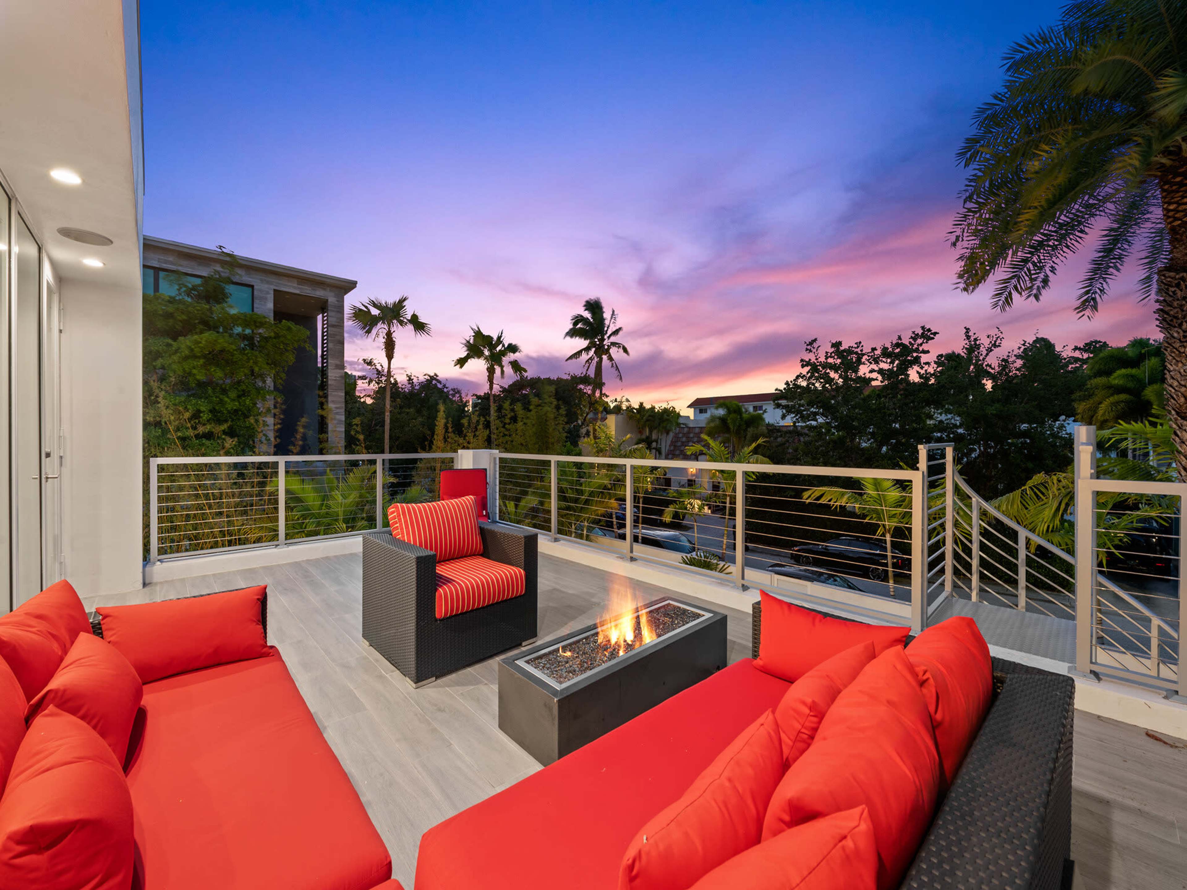A modern outdoor seating area features a fire pit surrounded by red cushions, with a backdrop of a colorful sunset and palm trees.