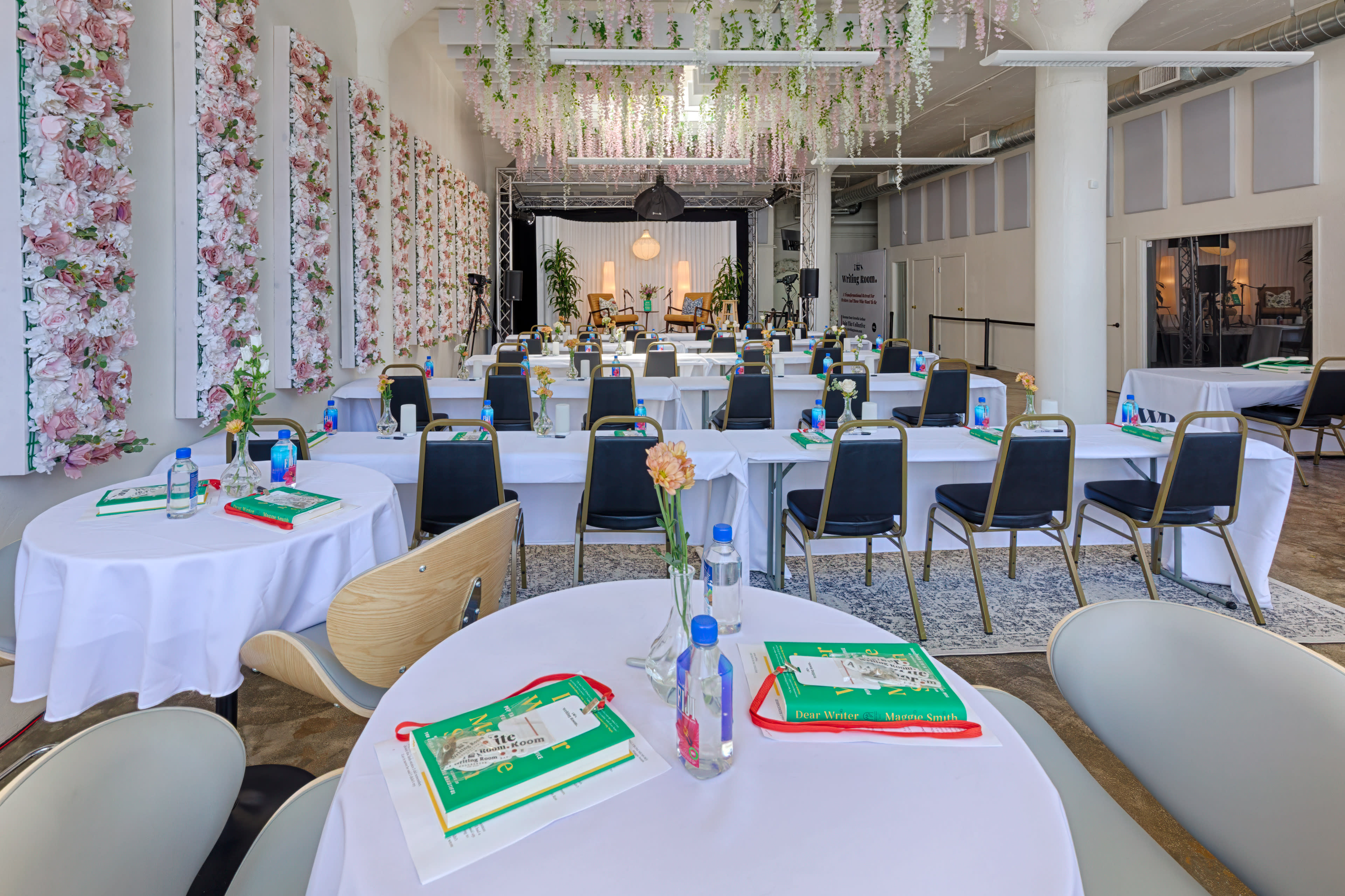 A spacious event room is arranged with round tables covered in white tablecloths, each featuring bottled water and notepads, with floral decorations adorning the walls and ceiling.