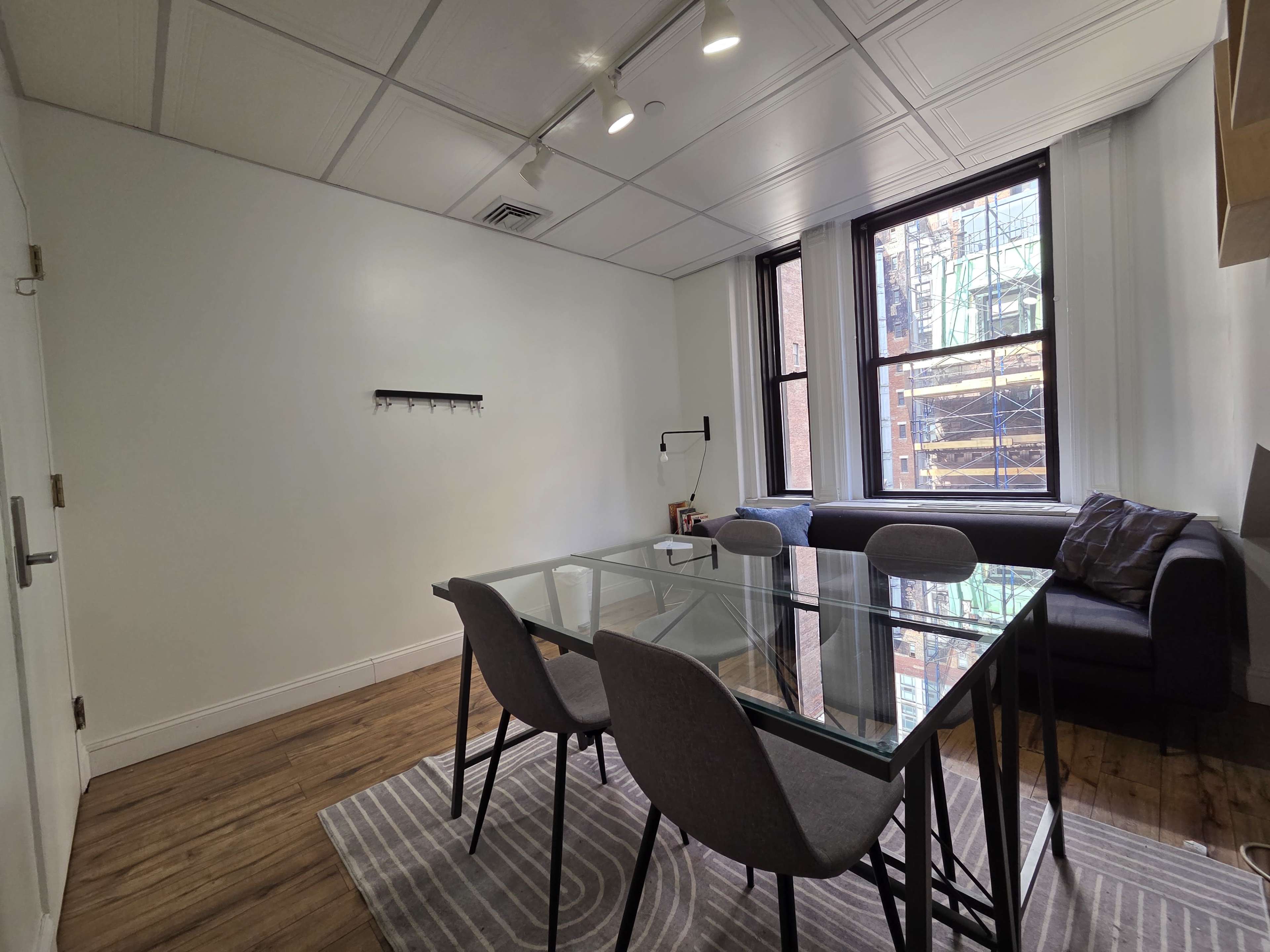 The image shows a modern office space with a glass dining table and gray chairs, a couch, and large windows revealing a view of a construction site outside.