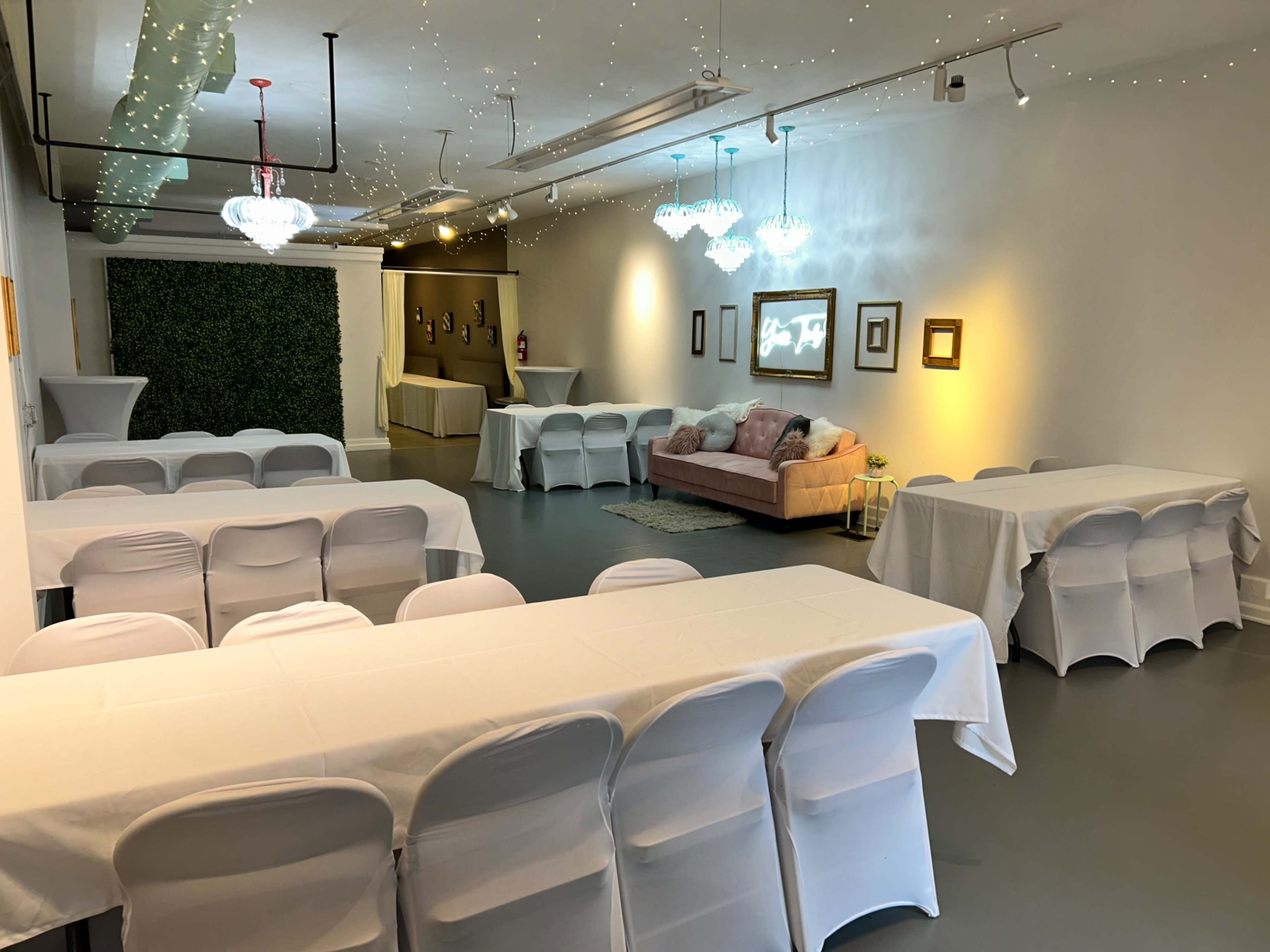 A spacious event room with neatly arranged tables covered in white tablecloths, a seating area with a sofa, and decorative lighting.