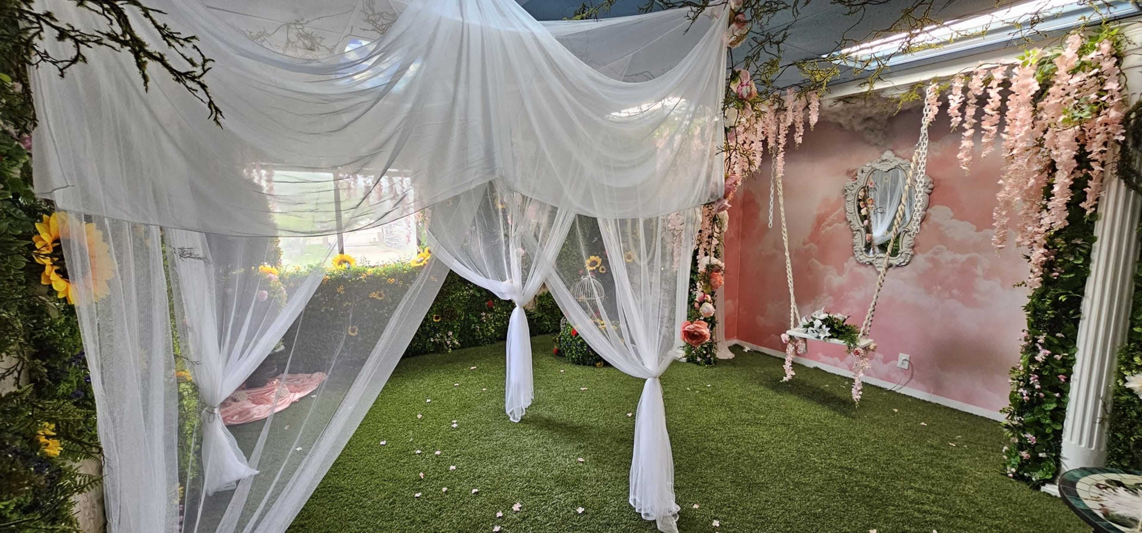 An elegantly decorated room featuring sheer white drapes, artificial grass, floral accents, and a mirror against a painted backdrop of soft clouds.
