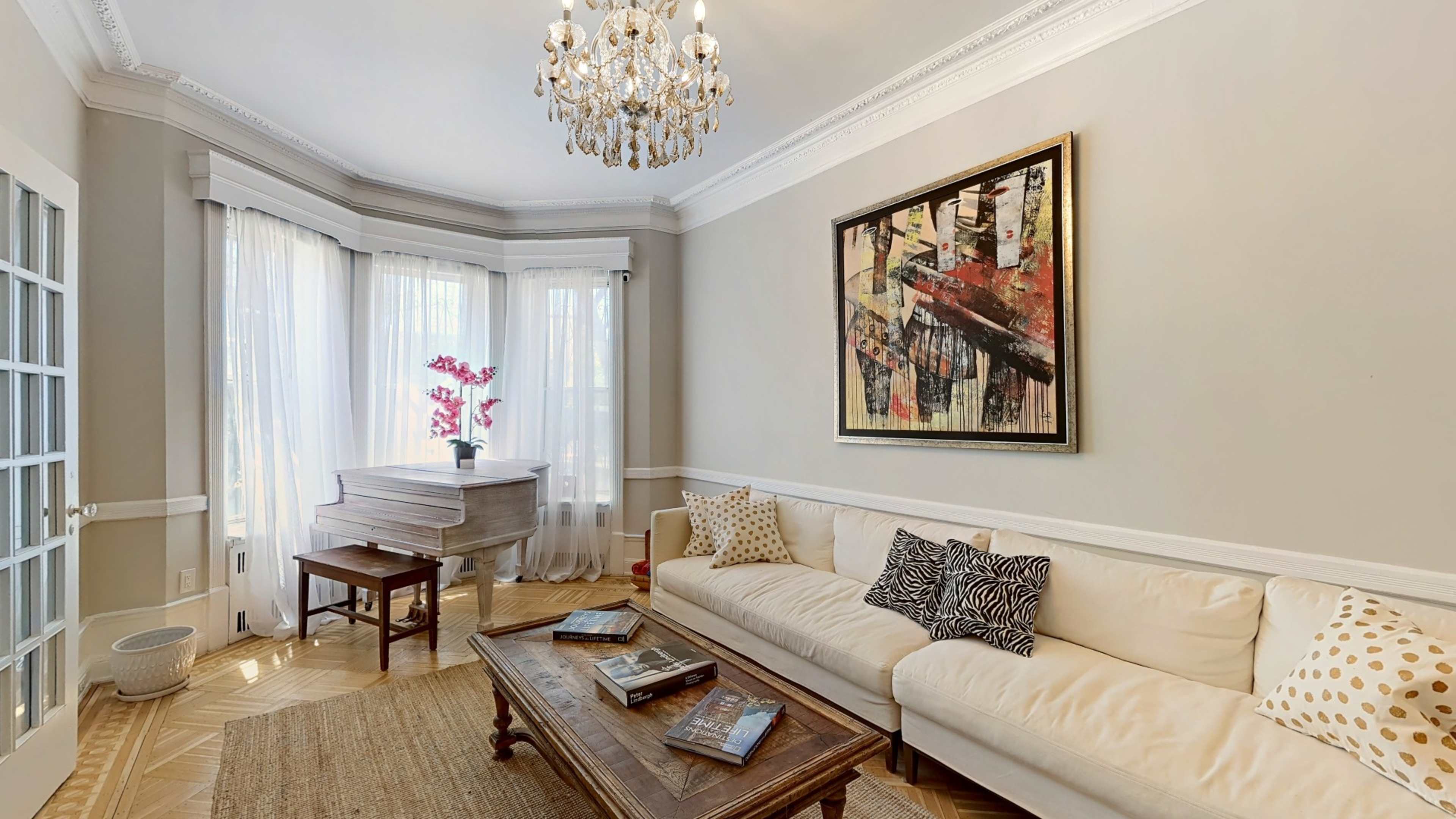 A well-decorated living room featuring a cream-colored sofa, an ornate chandelier, a painting on the wall, and a grand piano near a window with sunlight filtering through sheer curtains.