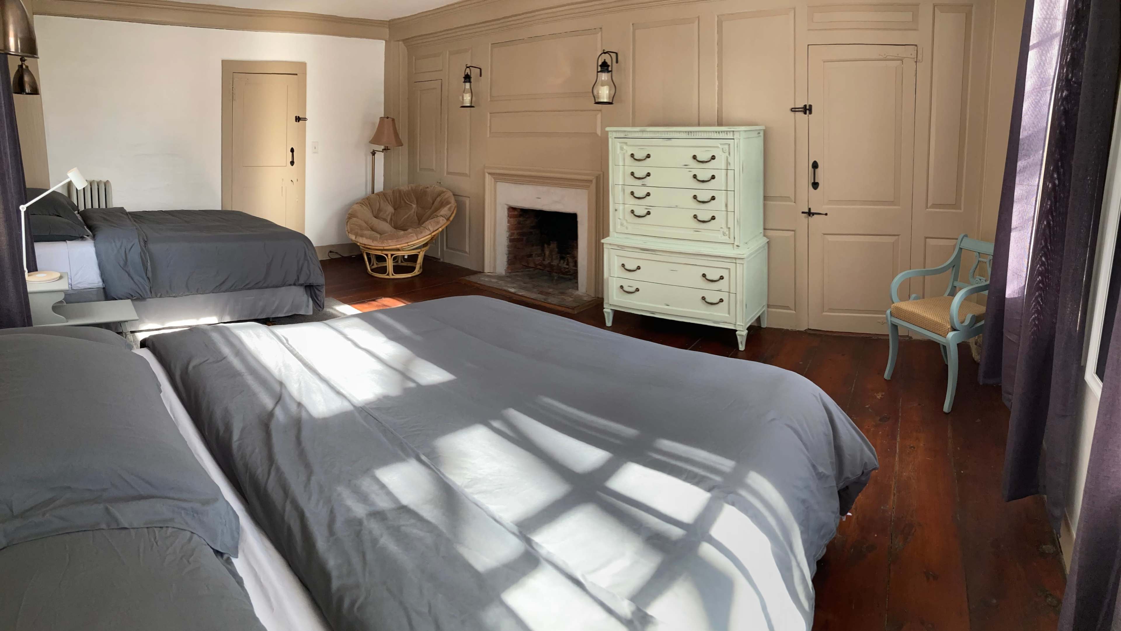 The image shows a bedroom with two large beds, a light green dresser, a chair, and a fireplace, illuminated by sunlight casting shadows across the wooden floor.
