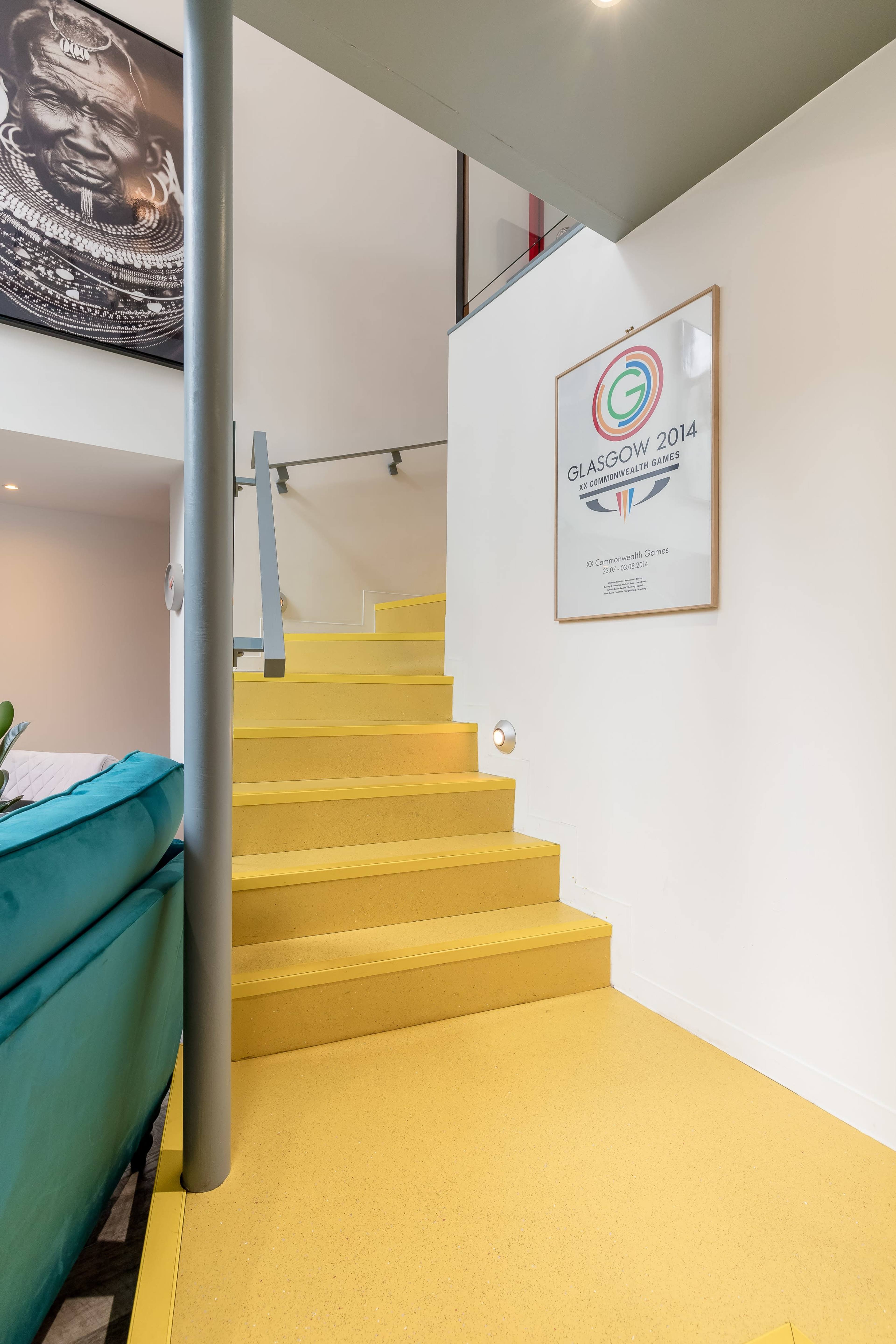 A brightly lit interior with a set of yellow stairs leading upward, accompanied by a framed poster on the wall.