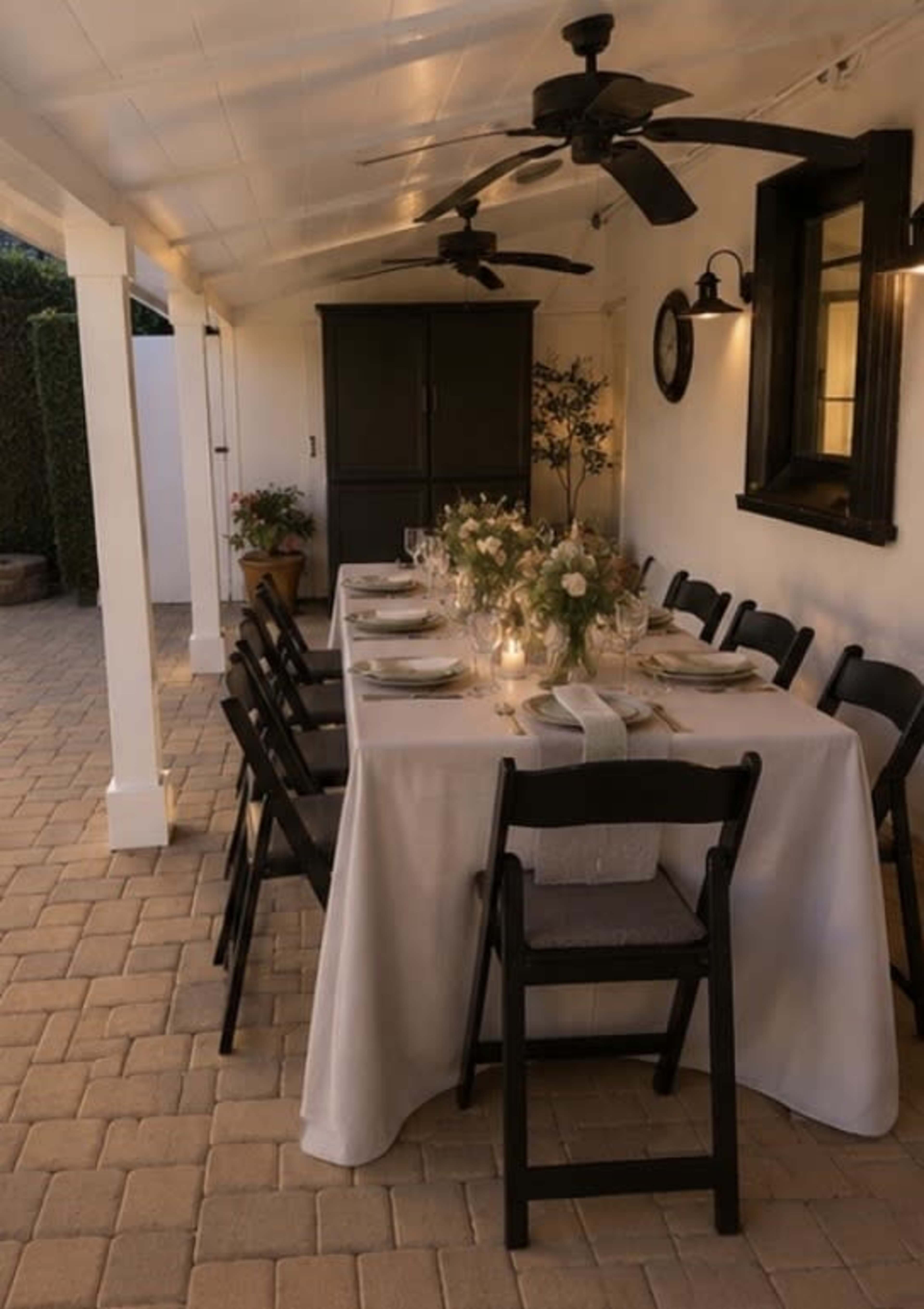A dining table is set outdoors under a covered patio, featuring a white tablecloth, floral centerpiece, and black chairs.