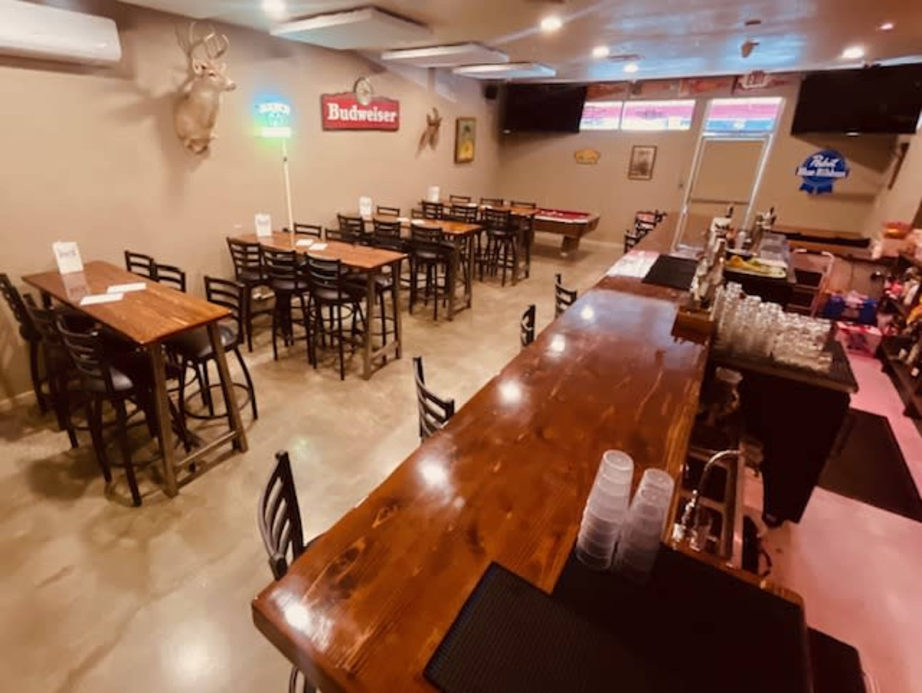 The image shows a spacious bar area with wooden tables and chairs, a long wooden bar counter, and various signs on the walls, including beer advertisements.