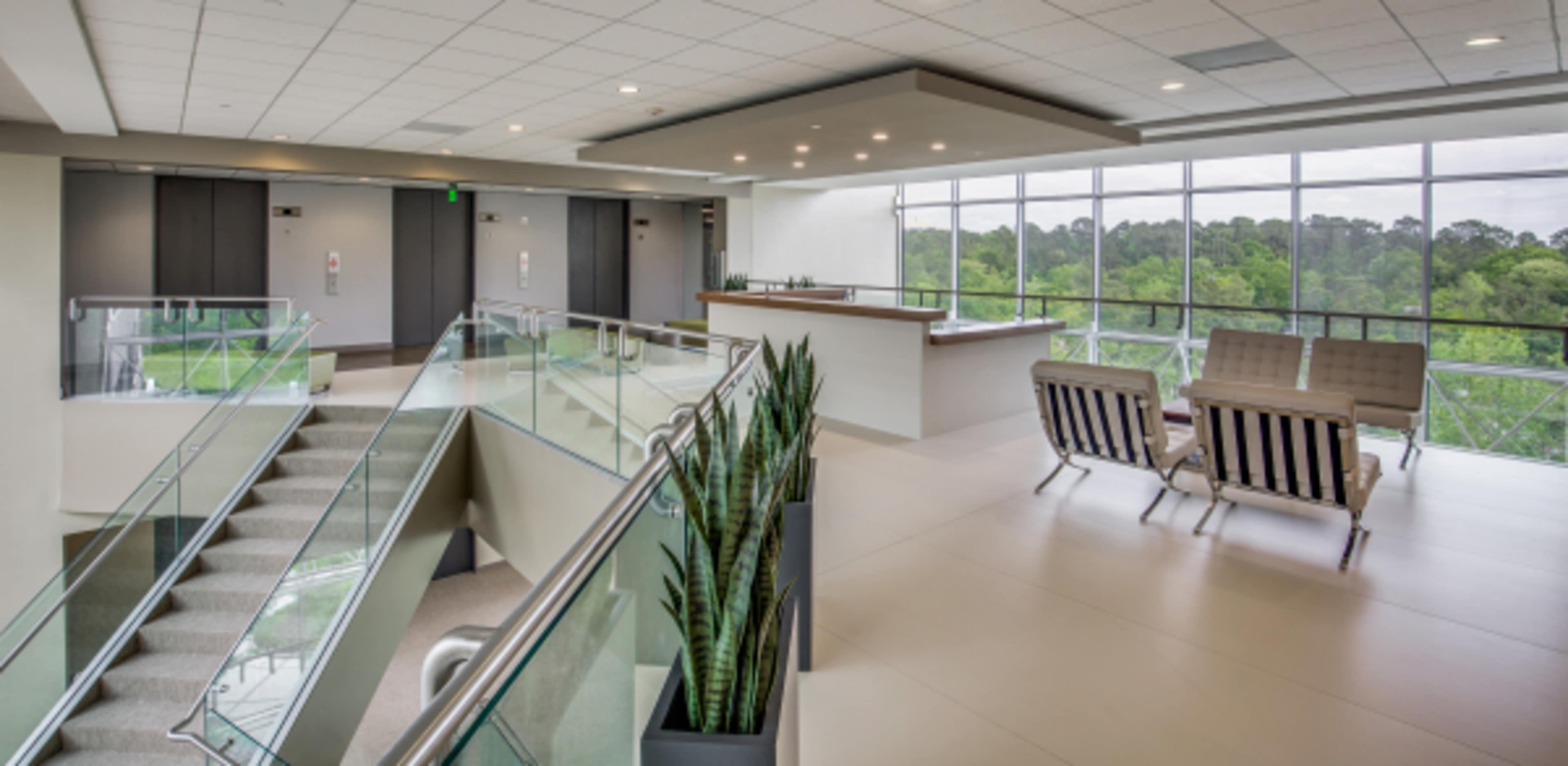 A modern lobby area featuring a staircase, a reception desk, and seating beside large windows with views of greenery outside.