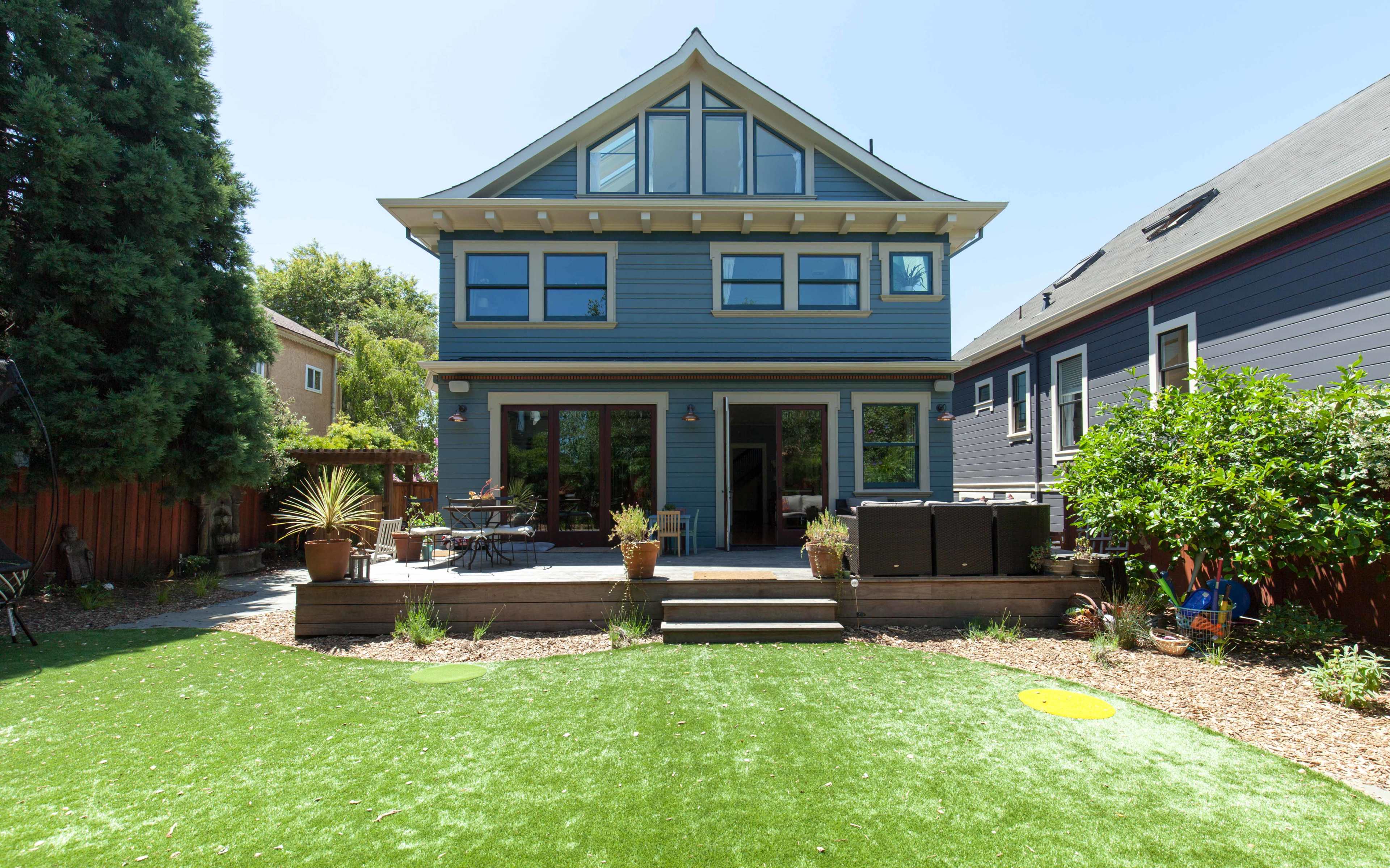 A two-story blue house with large windows and a landscaped backyard featuring artificial grass and patio furniture.