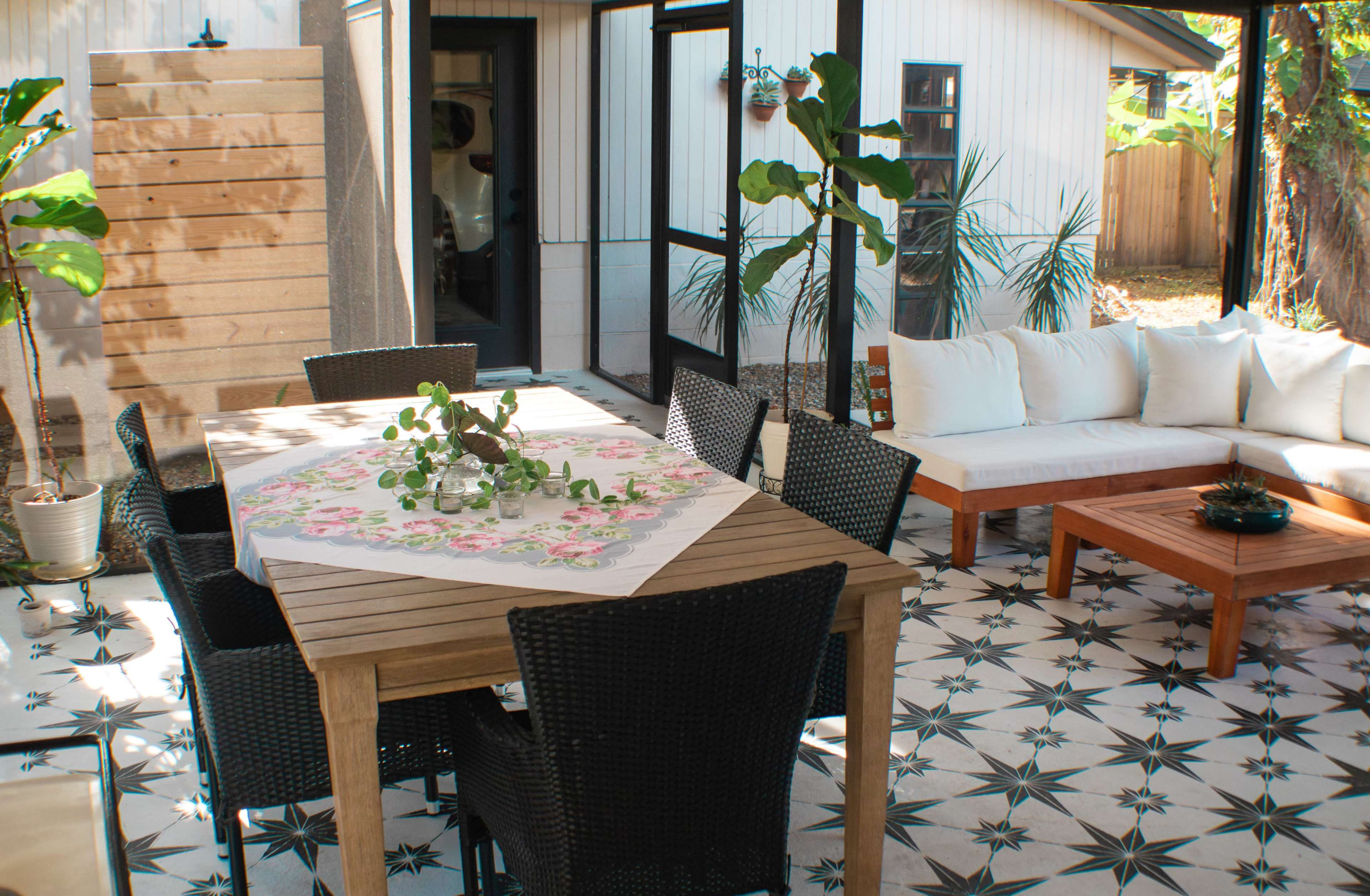 A modern outdoor patio with a wooden table surrounded by black chairs, a white sectional sofa, and patterned tile flooring.