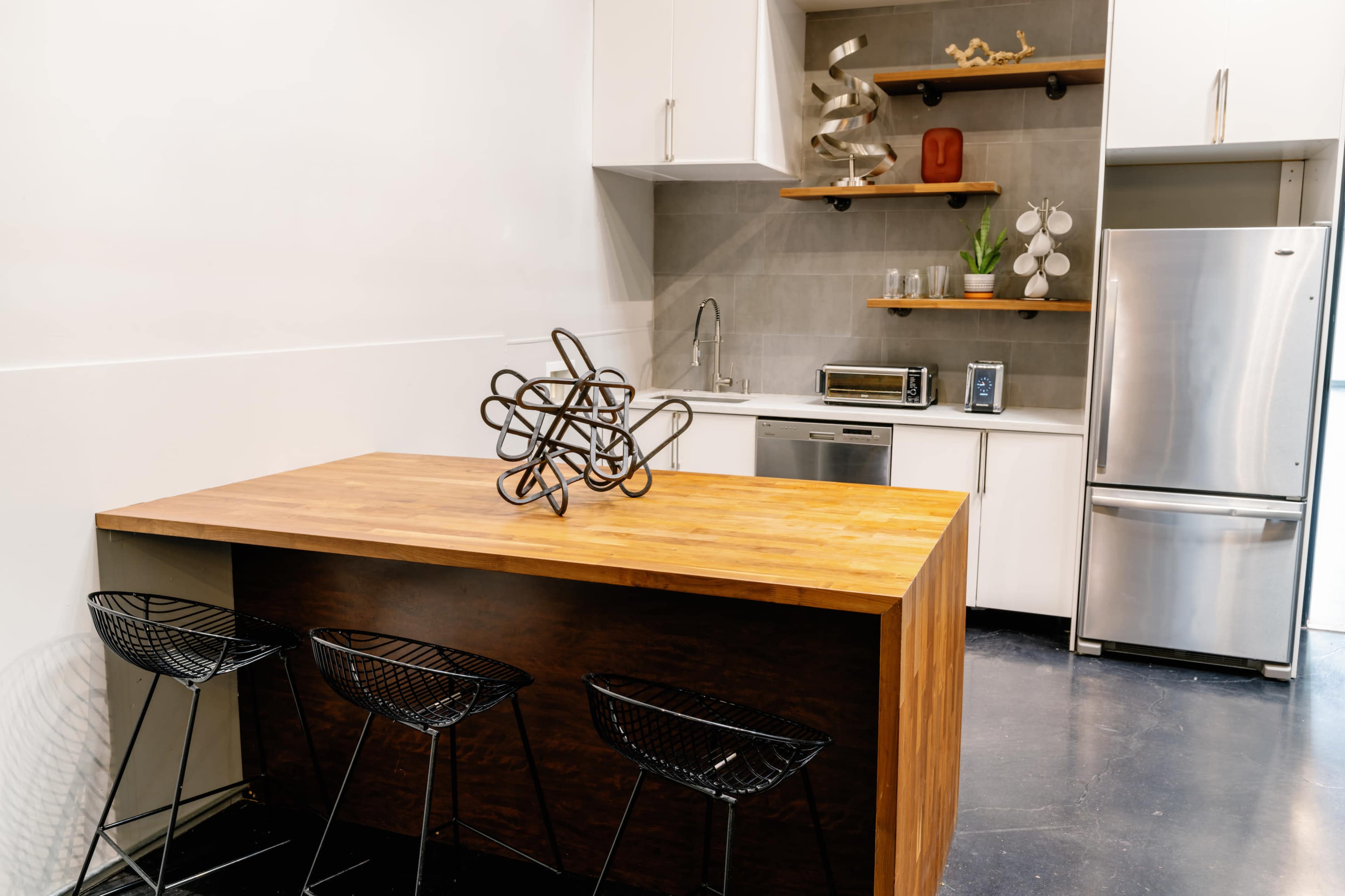 A kitchen features a wooden island with three black wire stools and a stainless steel refrigerator along a wall with open shelves and modern appliances.