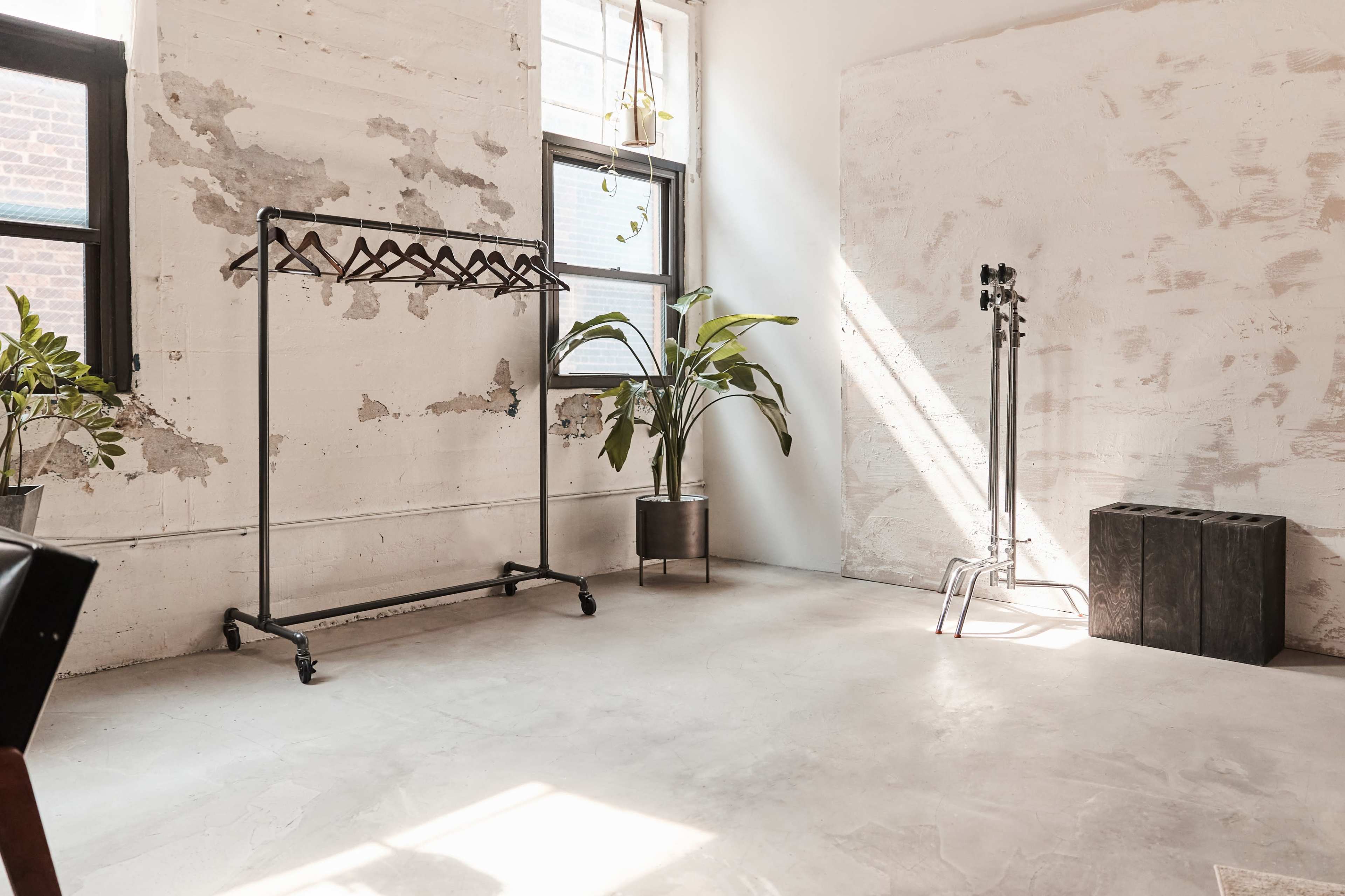 The image shows an empty room with exposed walls, a clothing rack, a potted plant, and a small black stool.
