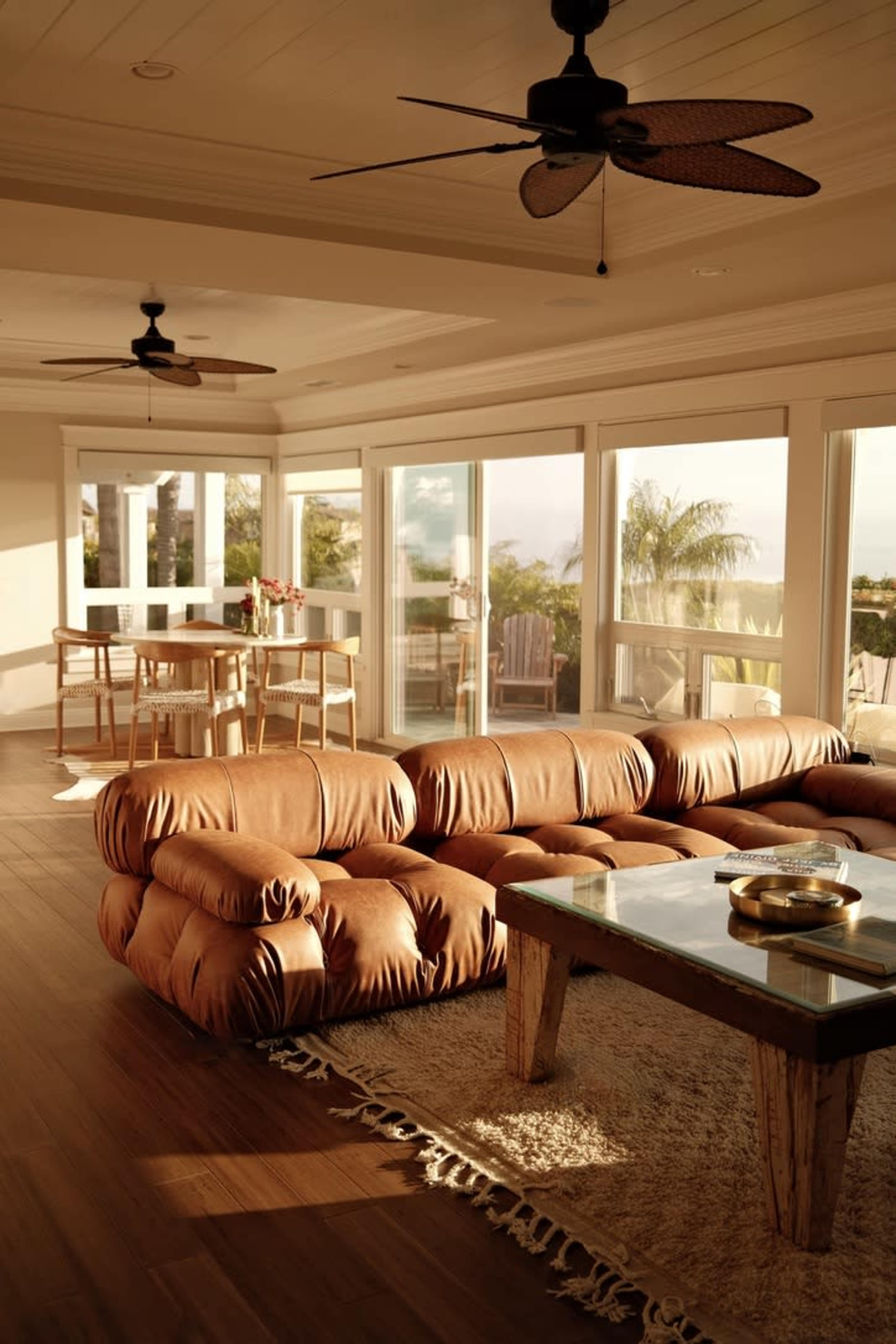 A spacious living room features a large brown sectional sofa, a wooden coffee table, and a dining area with tall chairs, illuminated by natural light from large windows.