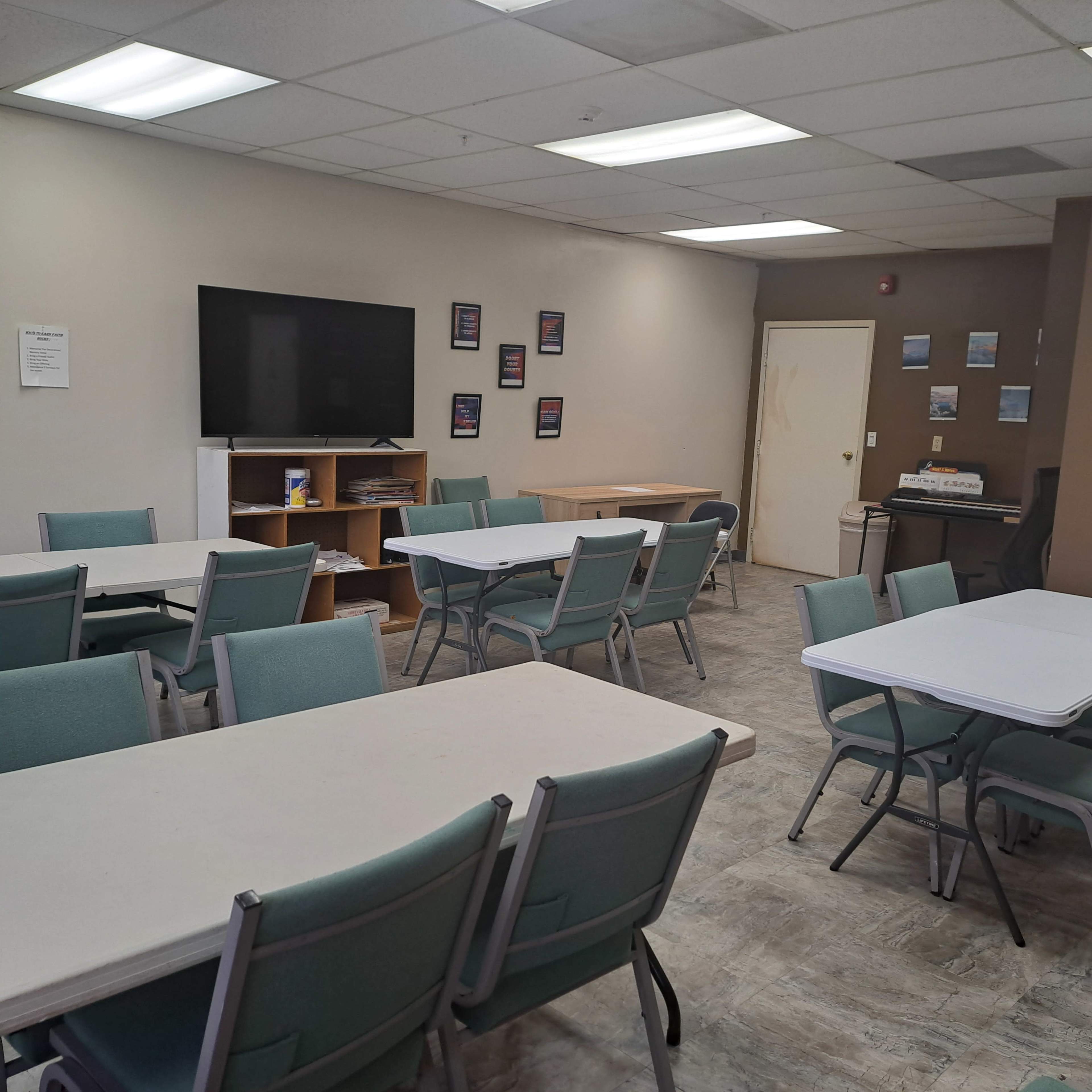 The image shows an empty community room with several tables and chairs arranged around a TV mounted on the wall, along with a door and a small workstation.