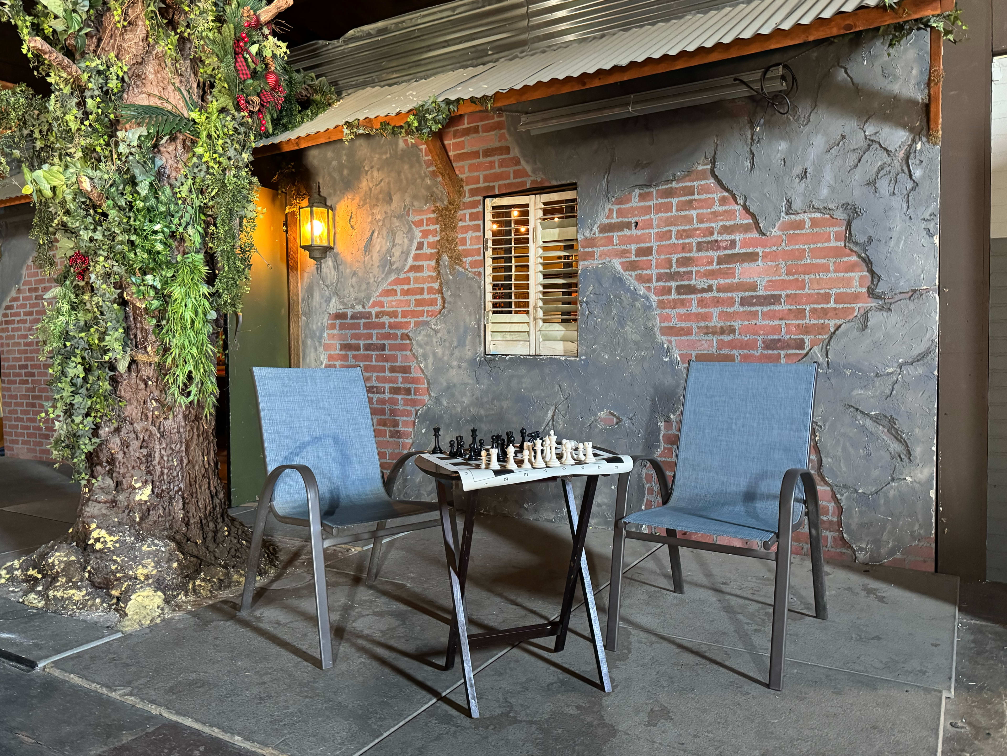A small bistro table with a chess set sits between two chairs on a patio surrounded by a brick wall and greenery.