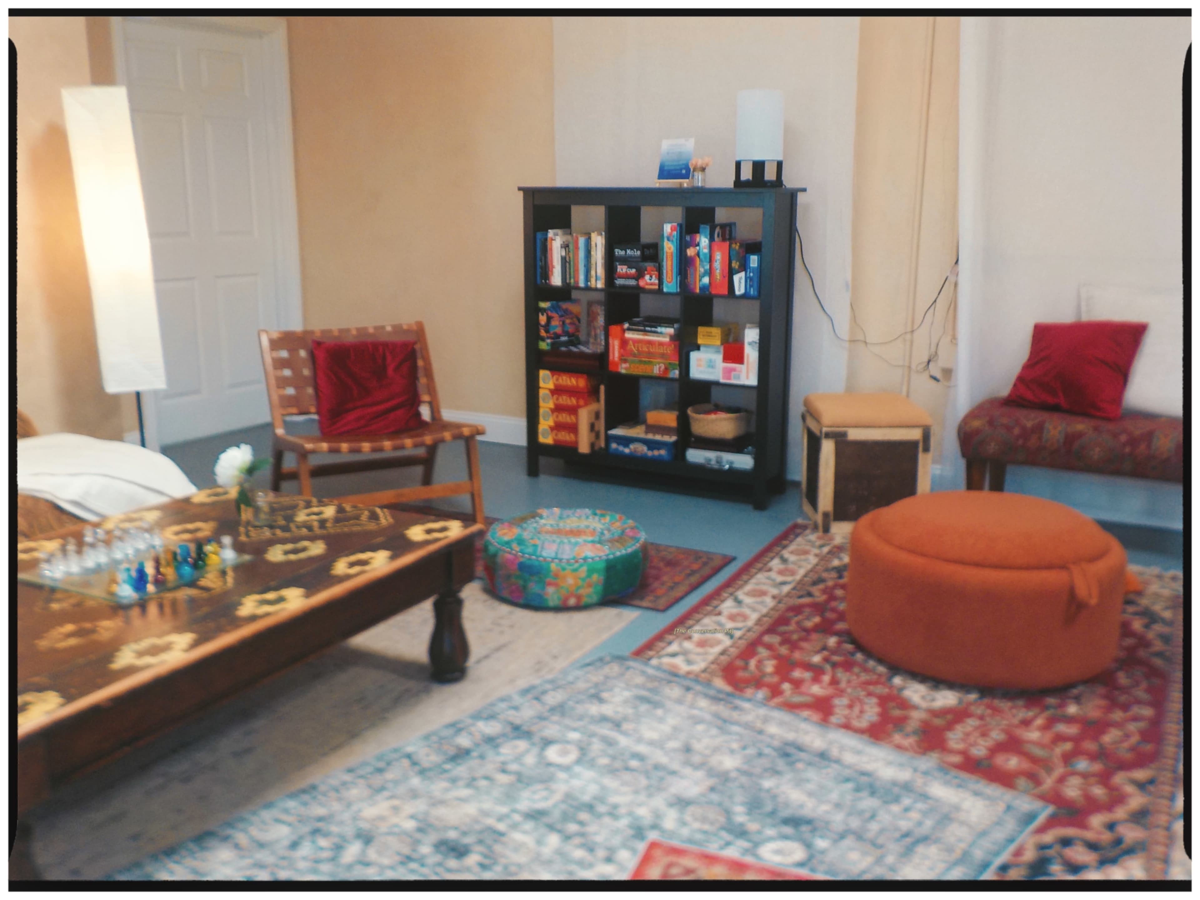 A cozy room features a wooden coffee table, a bookshelf filled with games, various seating options, and rugs on a blue floor.